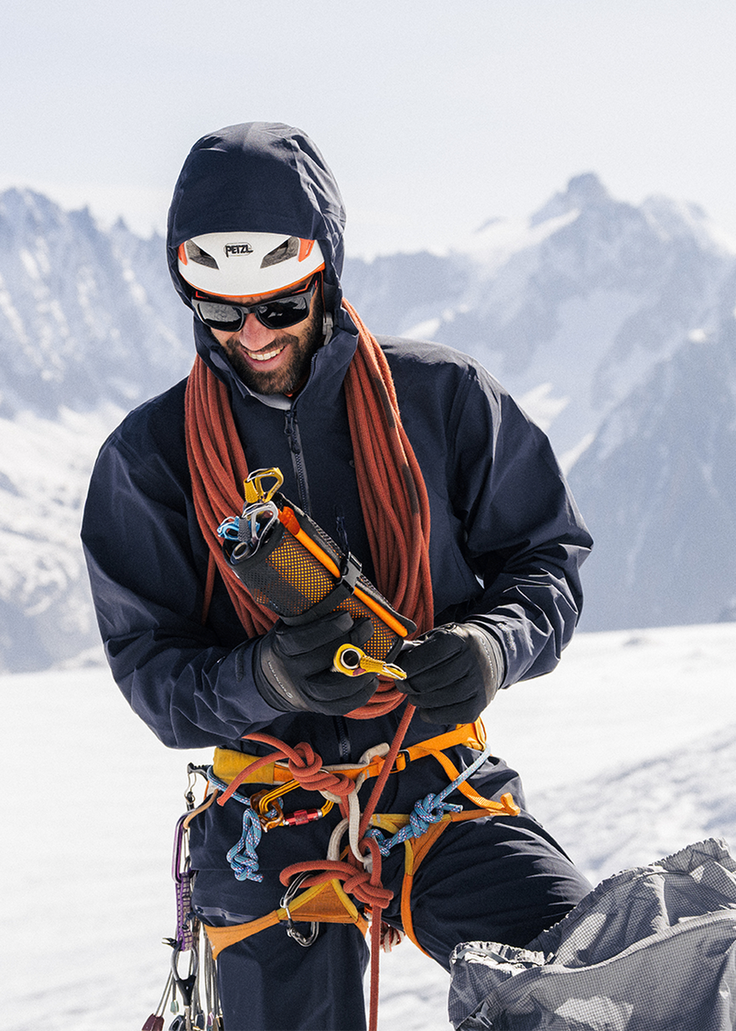 A climber in winter gear adjusts his equipment on a snowy mountain, with a backdrop of jagged peaks under a clear sky.