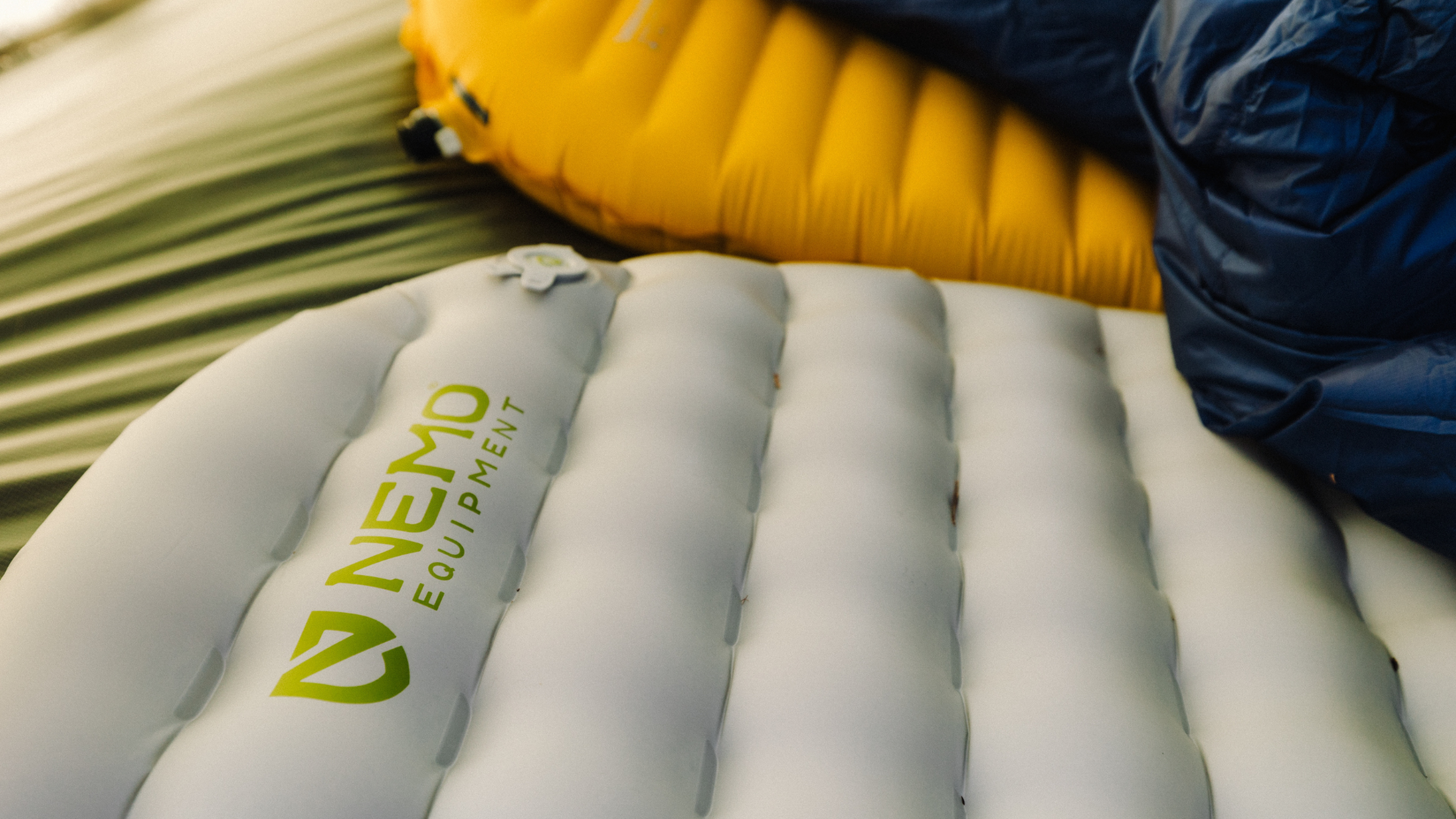 Close-up of a white NEMO Equipment inflatable sleeping pad with a yellow pad and blue sleeping bag in the background.