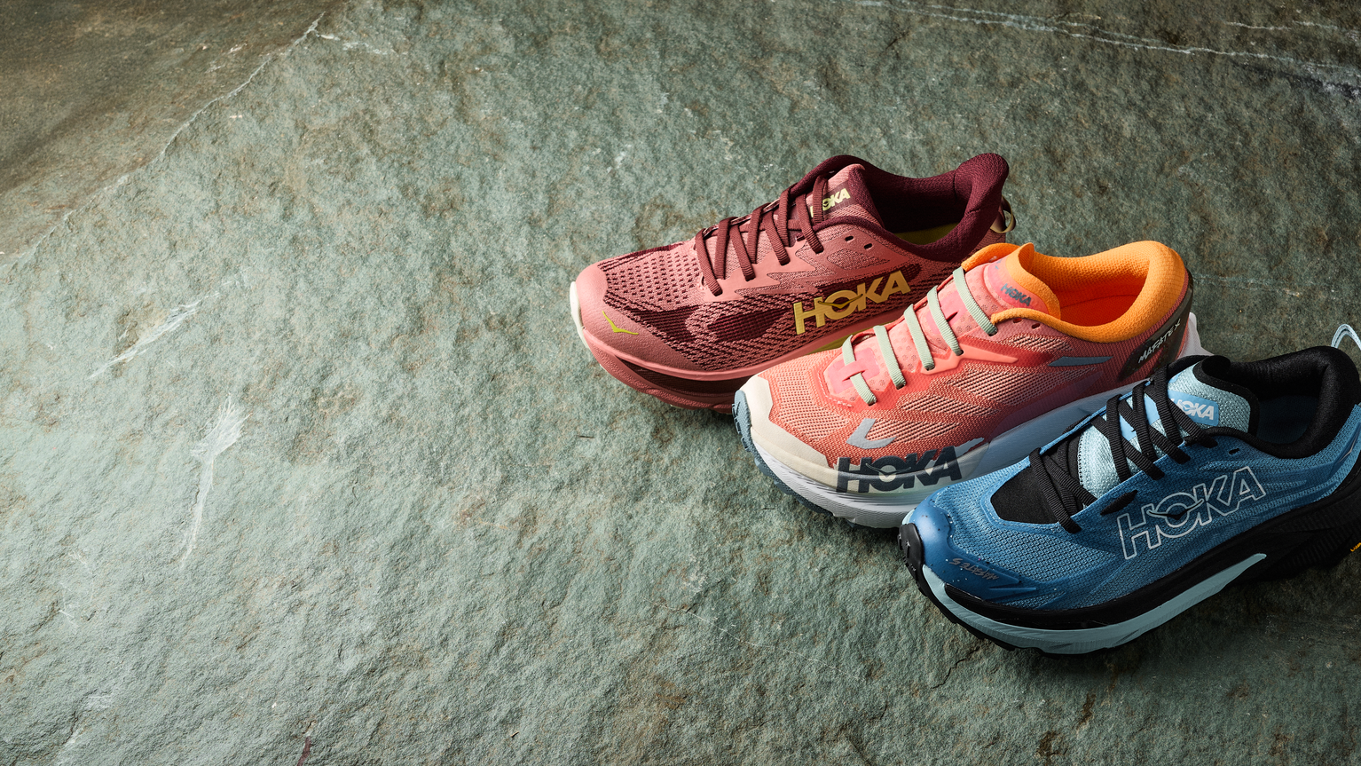 Three Hoka running shoes in pink, orange, and blue displayed on a textured stone surface, showcasing their design and colour variety.