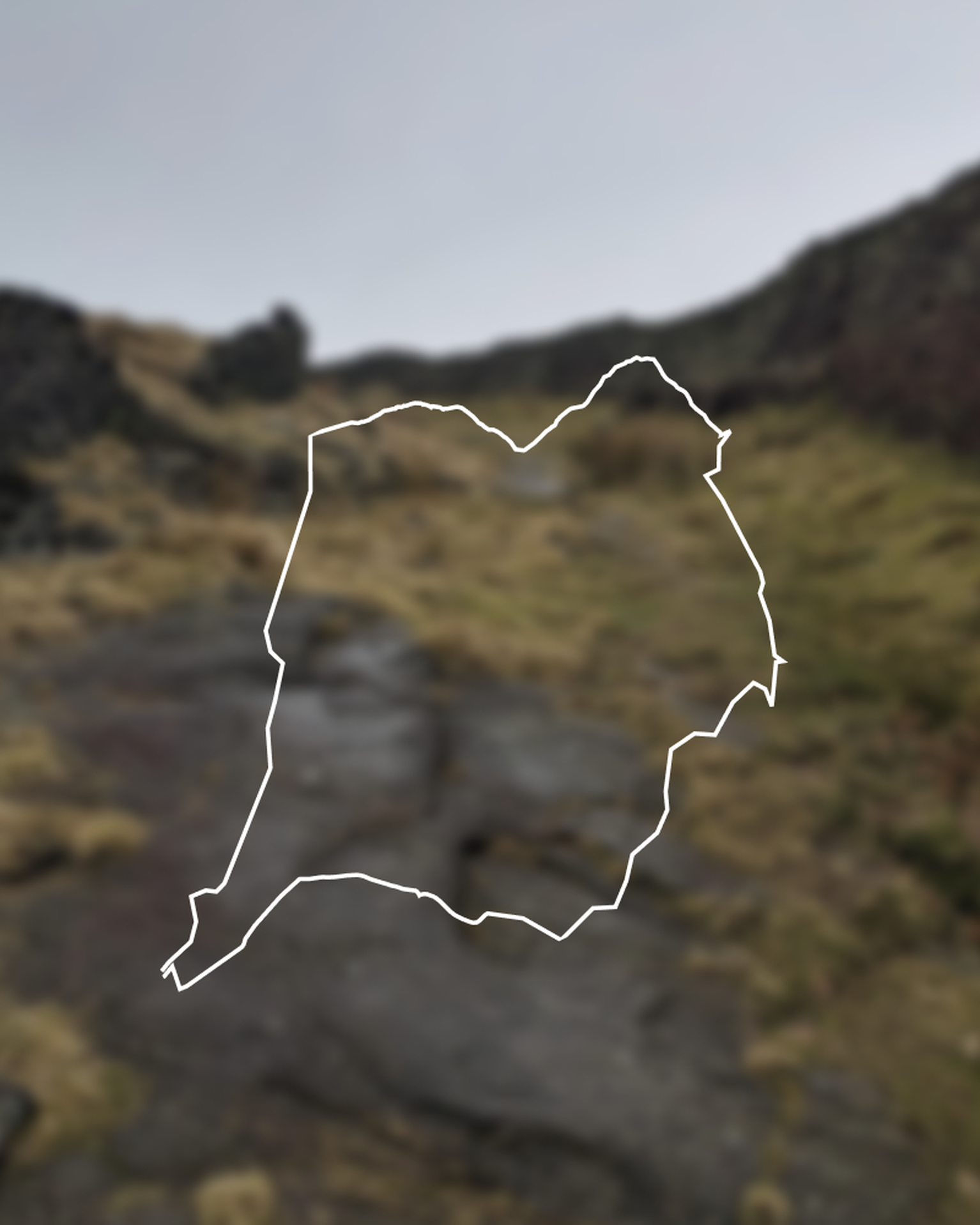 A blurred landscape with rocky terrain and grass, featuring a white outline of a walking route in the center.