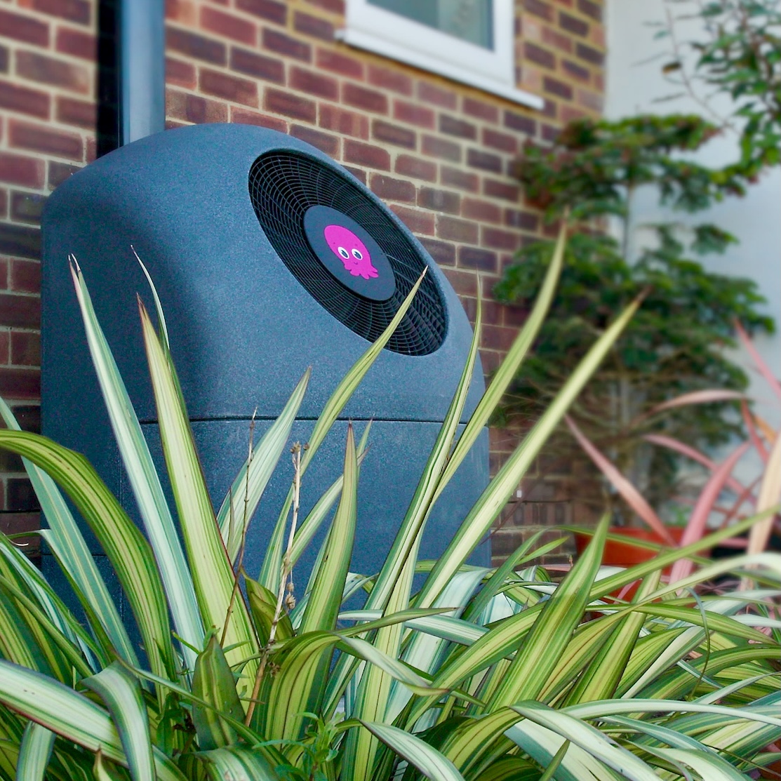 Outdoor Cosy heat pump with a fan featuring a pink octopus logo, surrounded by green spiky leaves, against a brick wall background.