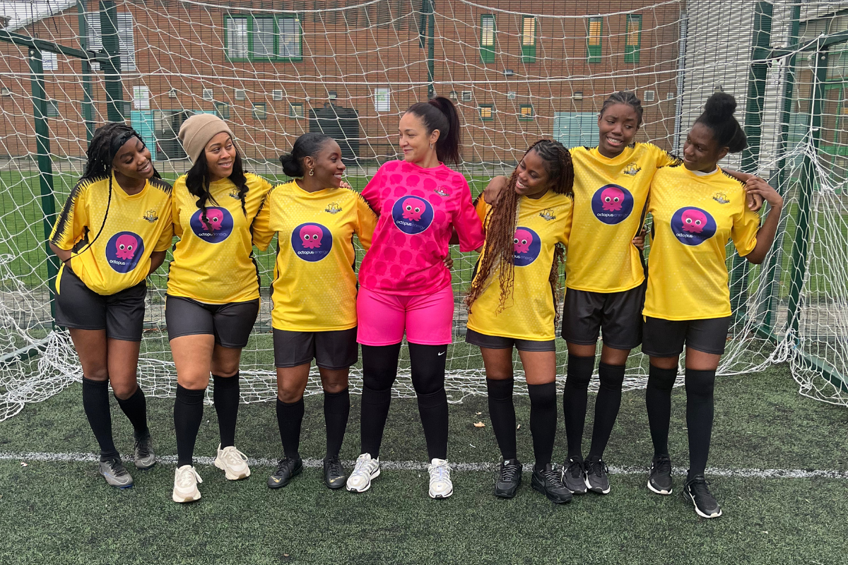 Seven female soccer players in yellow and pink uniforms stand arm-in-arm on the field, smiling in front of a goal net.