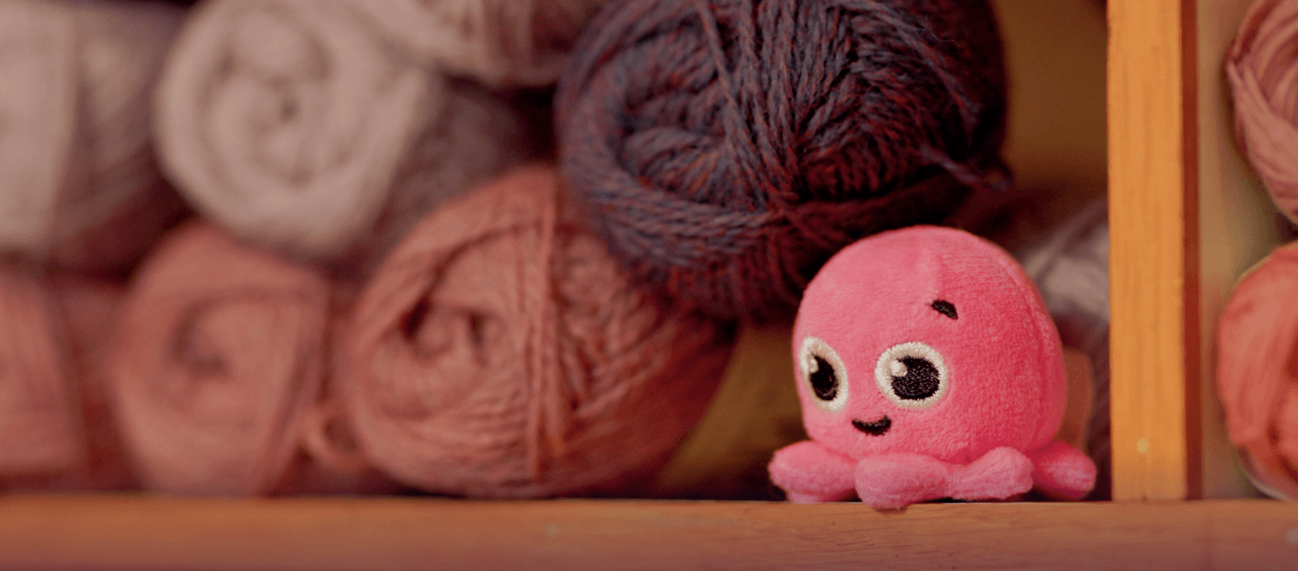 A cute pink octopus plush toy sits in front of a shelf filled with colorful yarn balls.