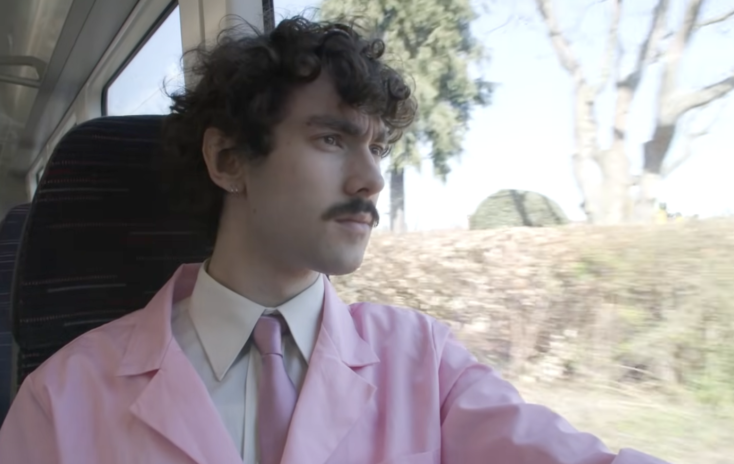 A person with curly hair and a mustache gazes out a train window, wearing a pink suit and tie. Trees and bushes blur past outside.