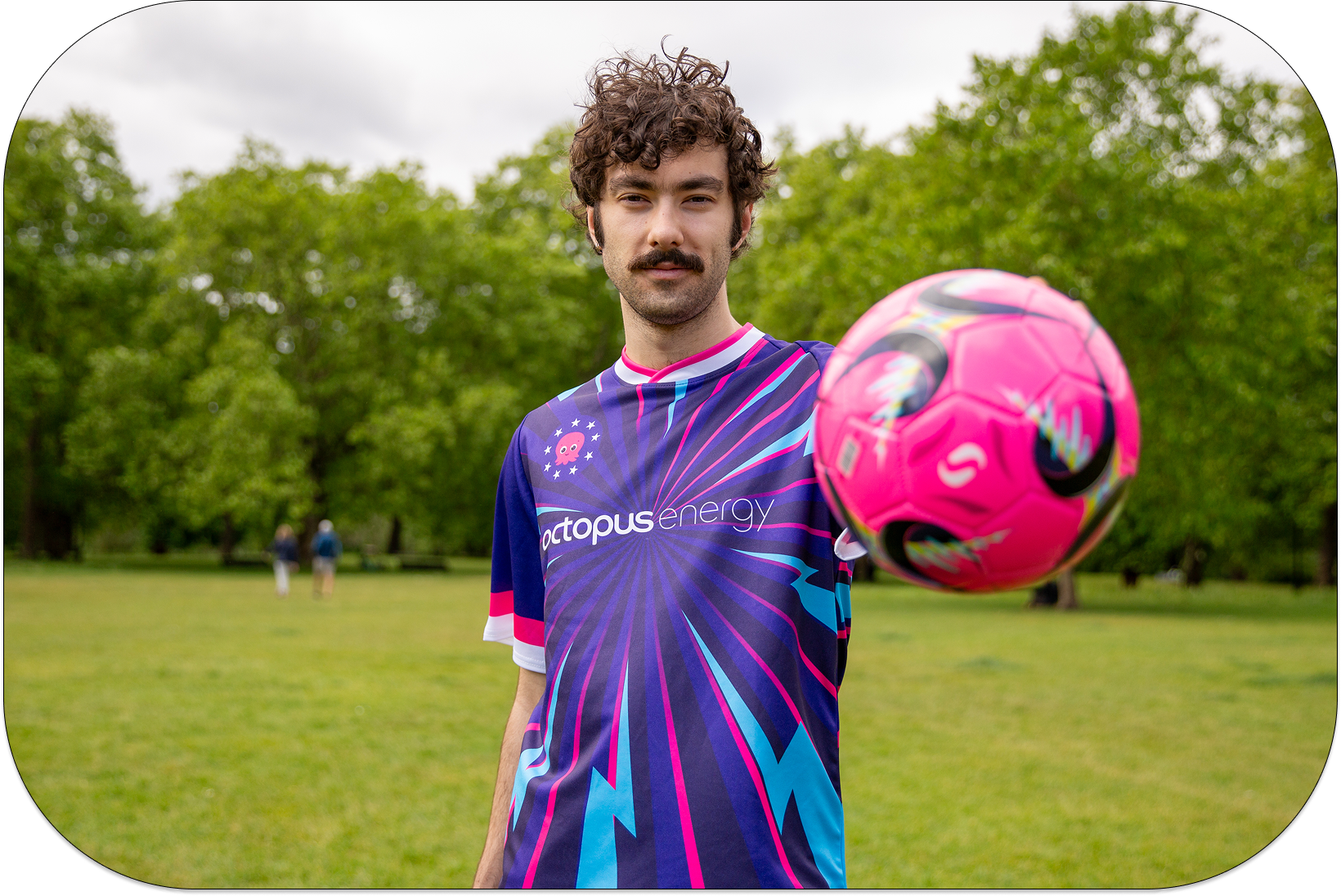 Man in a colorful Octopus Energy jersey holding a pink soccer ball in a park, with trees in the background.