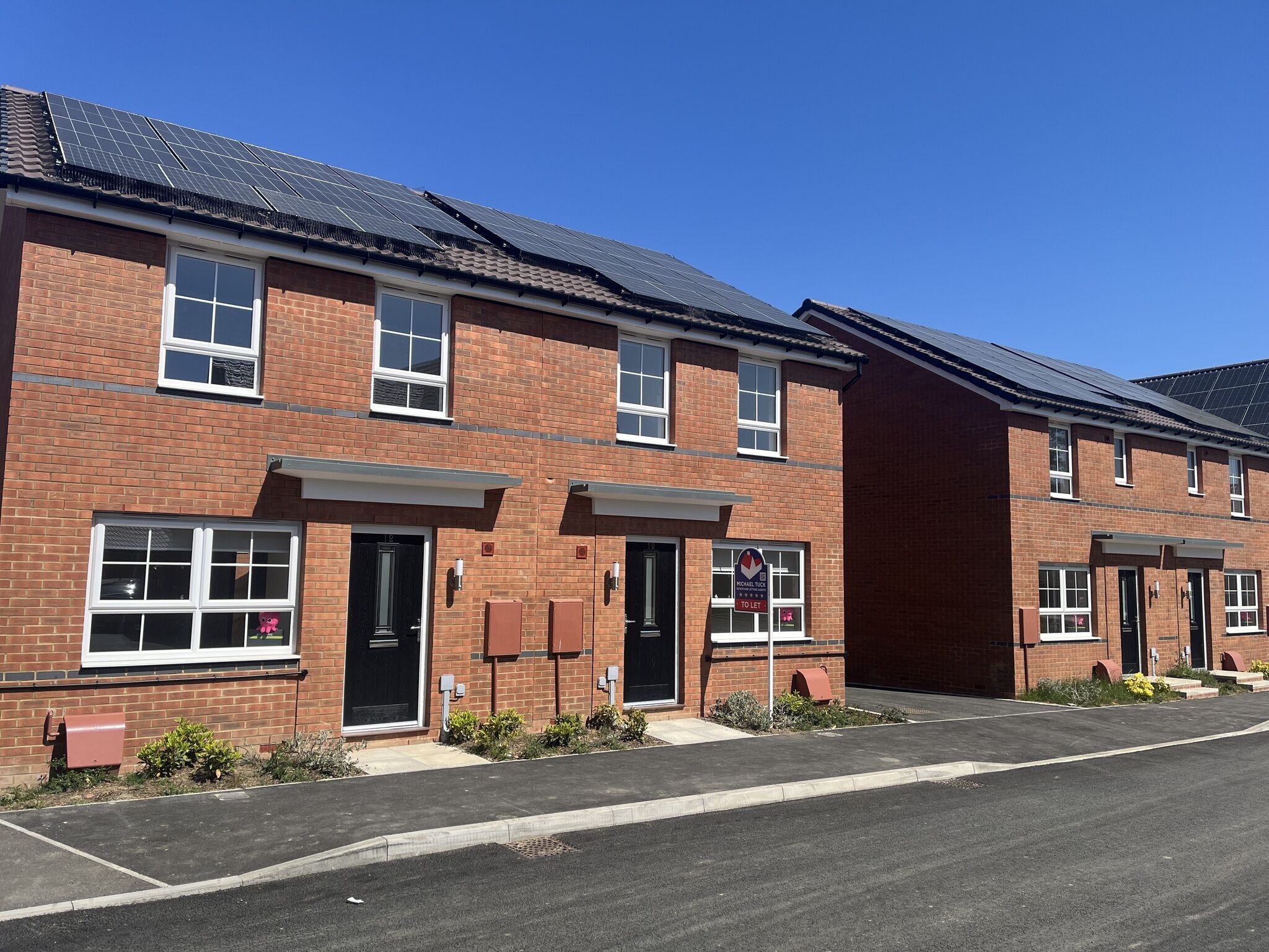 Red brick semi-detached houses with solar panels under a clear blue sky. A "To Let" sign is visible in front of one home.