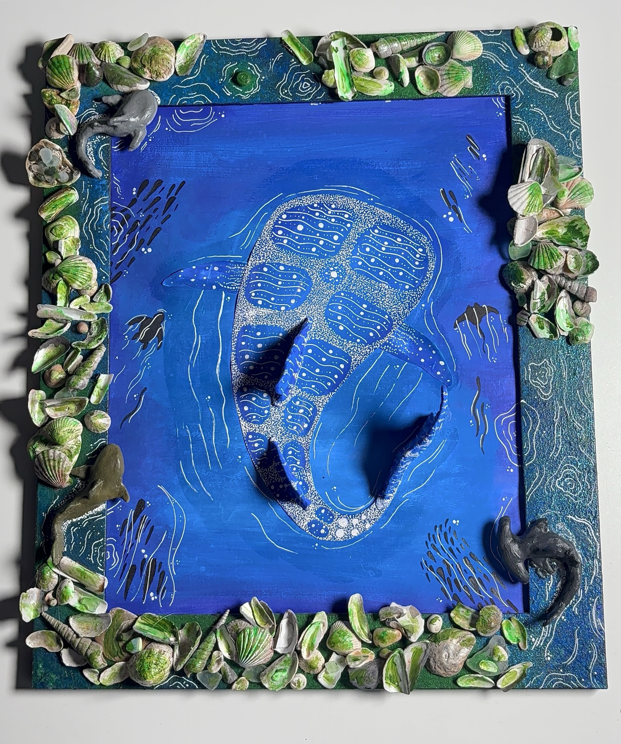 Painting of a manta ray with intricate patterns swimming against a blue background, bordered by seashells and tiny sculpted marine creatures.