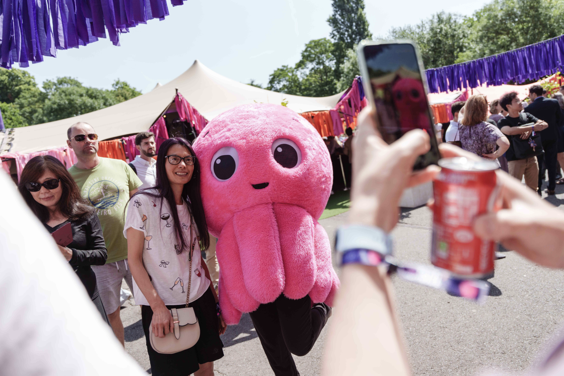 Person posing with a pink octopus mascot at an outdoor event, while another takes a photo. Colorful streamers decorate the tents.