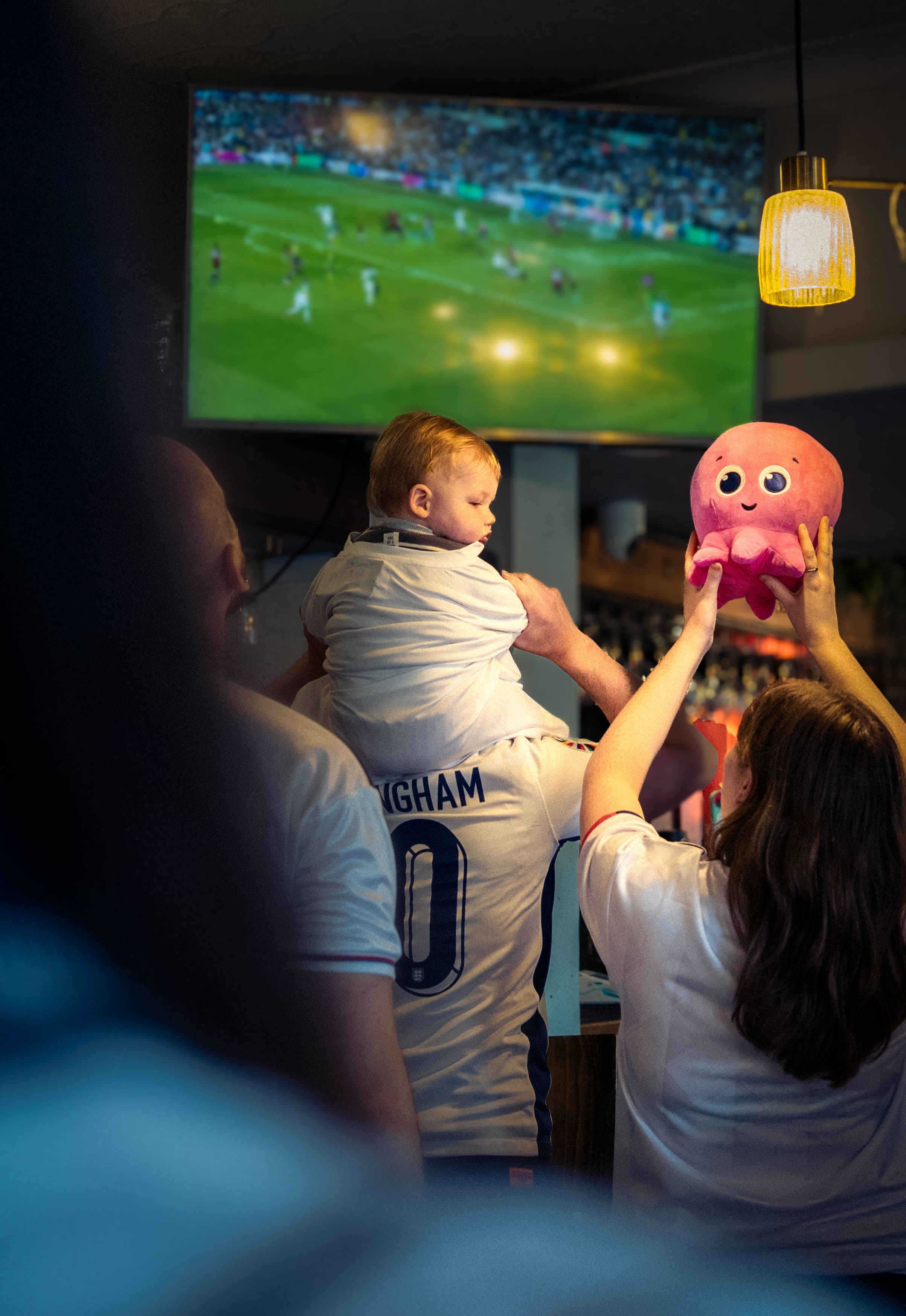A man holds a child on his shoulders, watching a football match on a TV as a woman lifts a pink toy octopus.