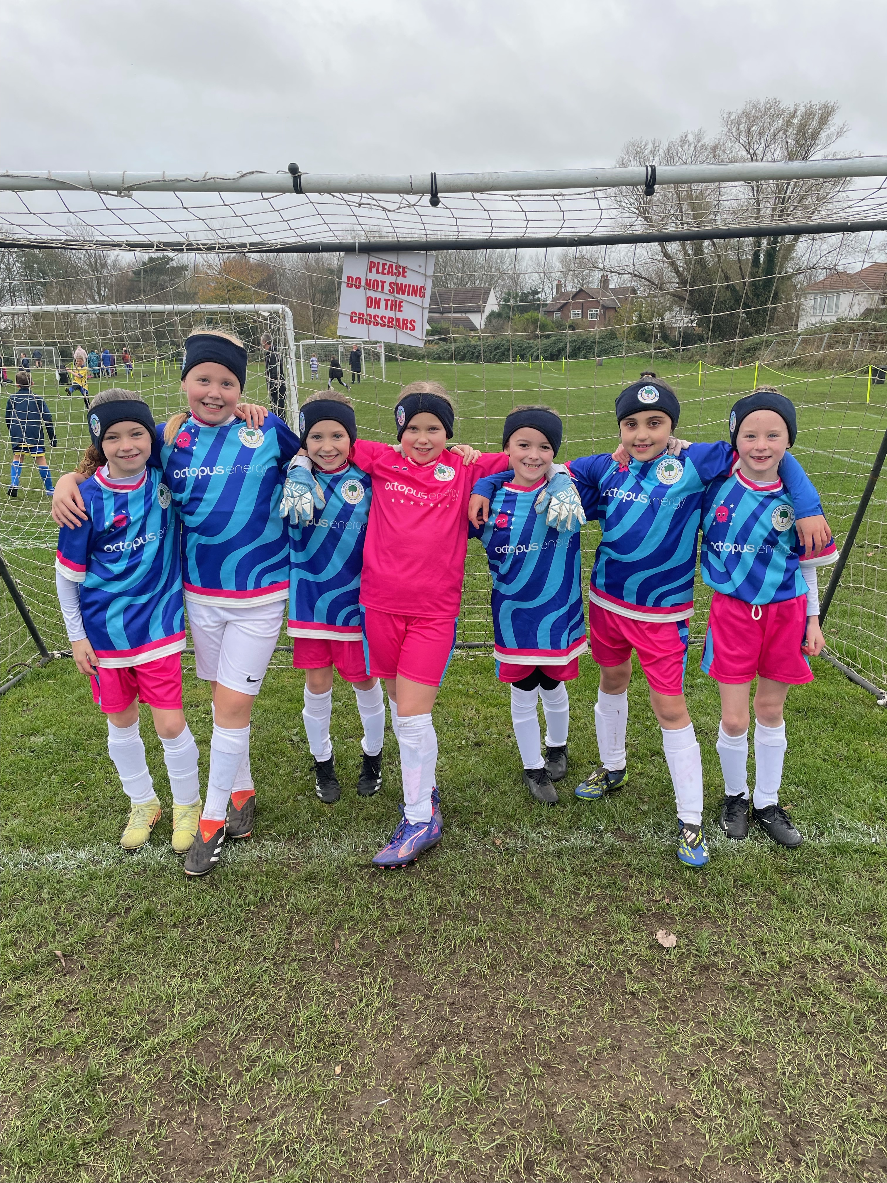 A group of young soccer players in blue and pink Octopus football kits stand smiling in front of a goal on a grass field, arms around each other.