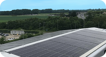 Solar panels installed on a rooftop with a scenic view of greenery, fields, and a road in the background on a clear day.