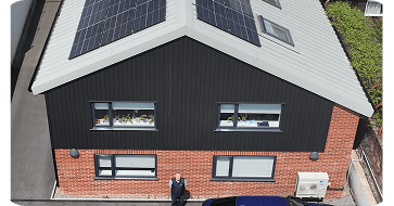 Aerial view of a two-story building with solar panels on the roof, red brick facade, and large windows. A person stands in front of the building.