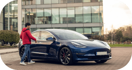 Man in a red jacket approaching a blue car parked in front of a modern glass building.