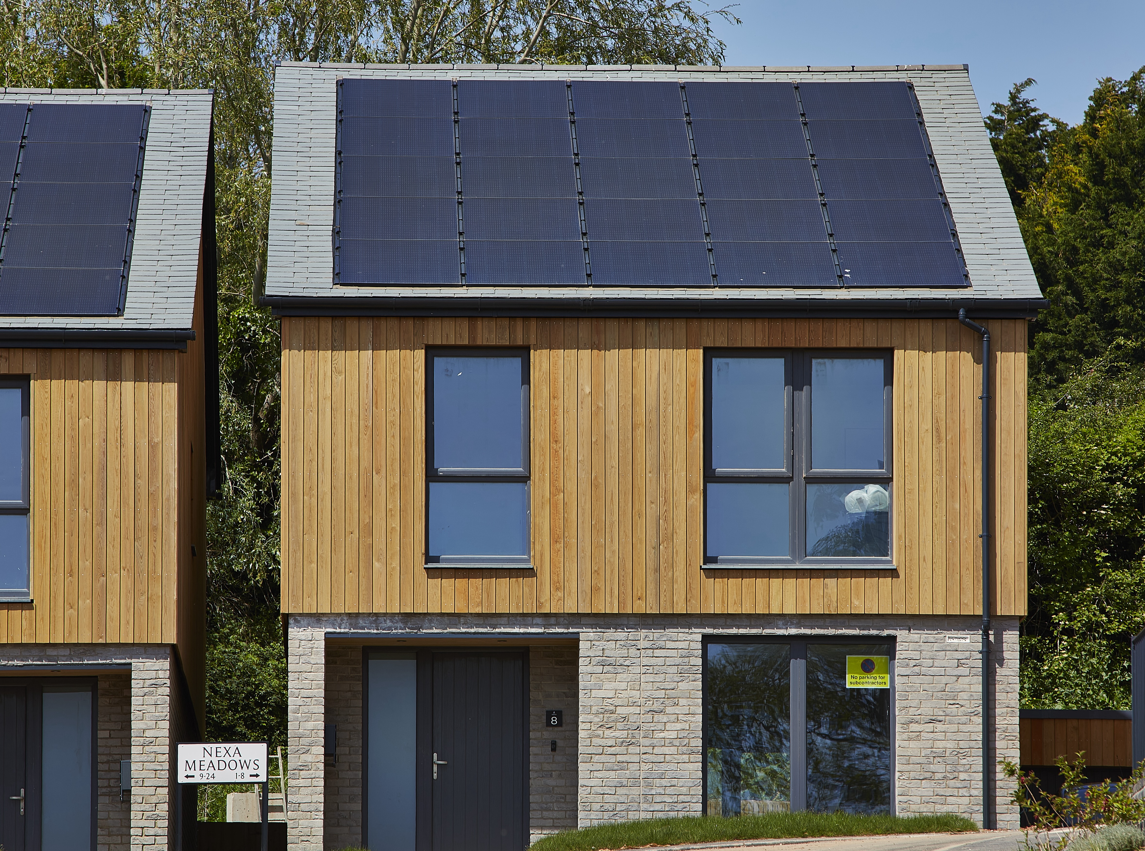 Modern two-story house with solar panels, wood cladding, and large windows. Sign reads "Nexa Meadows." Trees in the background.