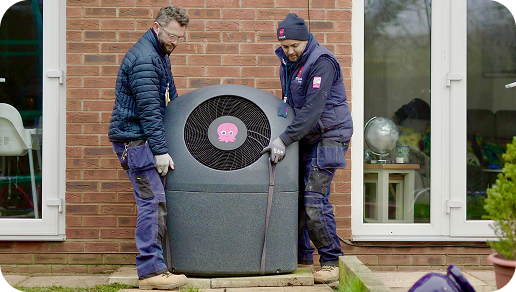 Two workers in jackets install a large heat pump unit outside a brick house with glass doors on a wooden platform.