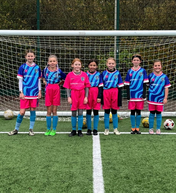 Six girls in matching soccer uniforms stand in front of a goal on a field, smiling and ready to play.