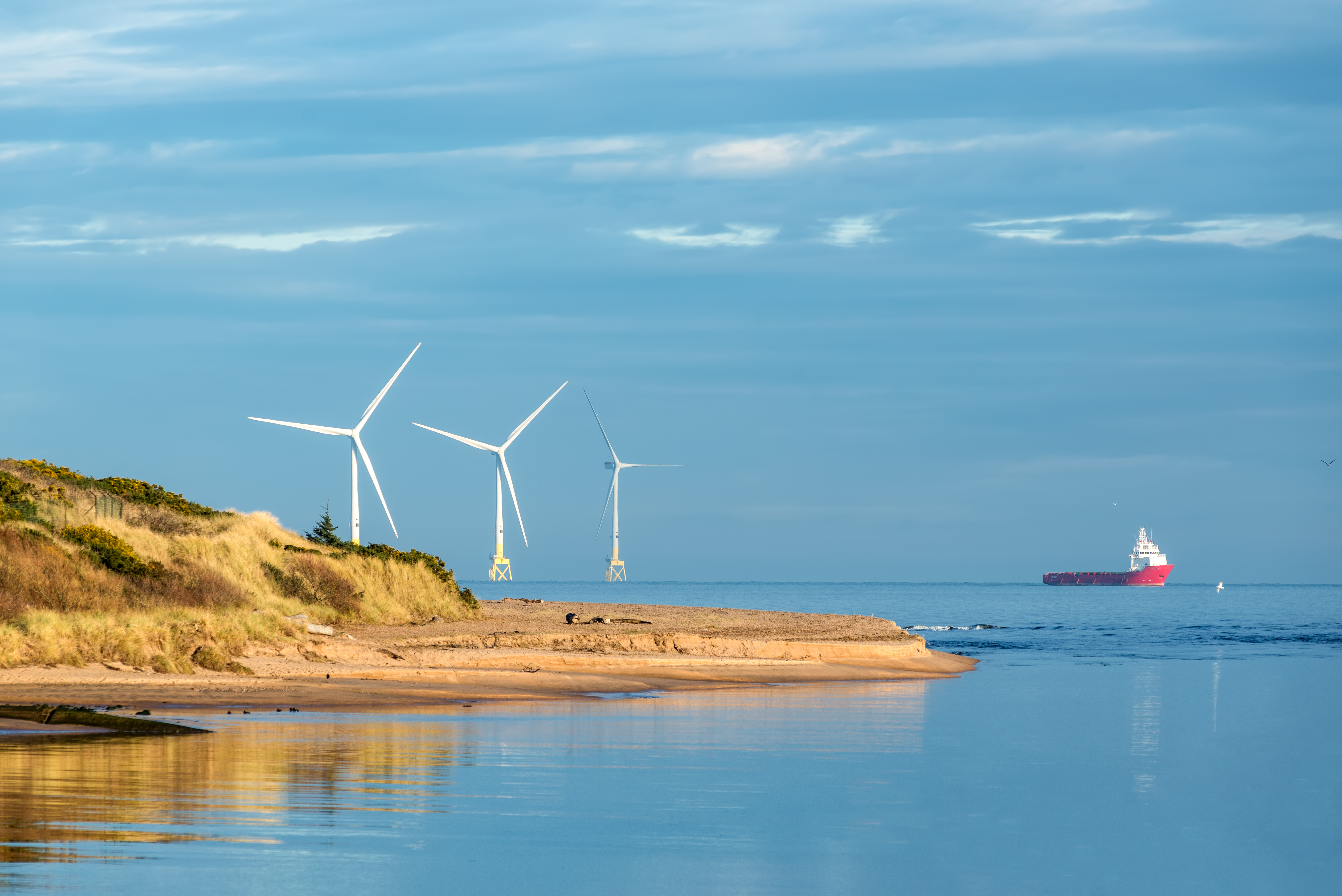Three wind turbines on a coastal beach with a red cargo ship in the distance under a blue sky.