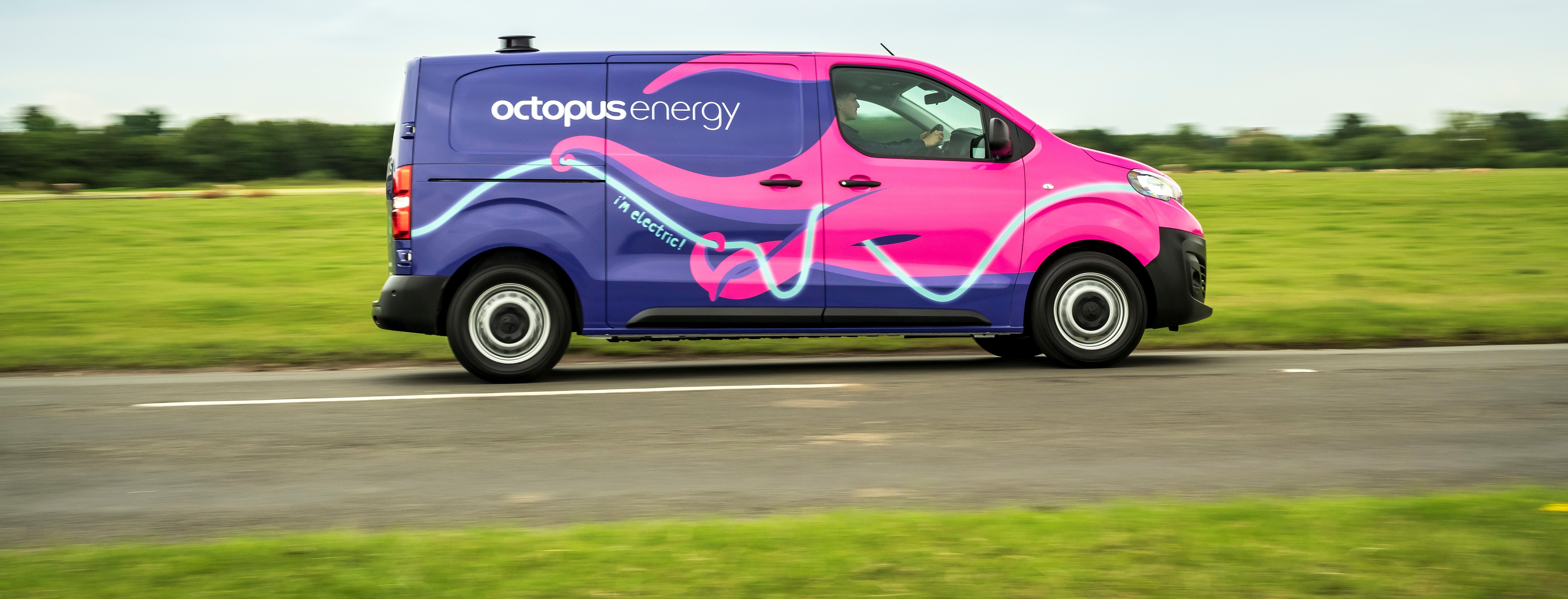 A purple and pink Octopus Energy electric van driving on a rural road with green fields in the background.