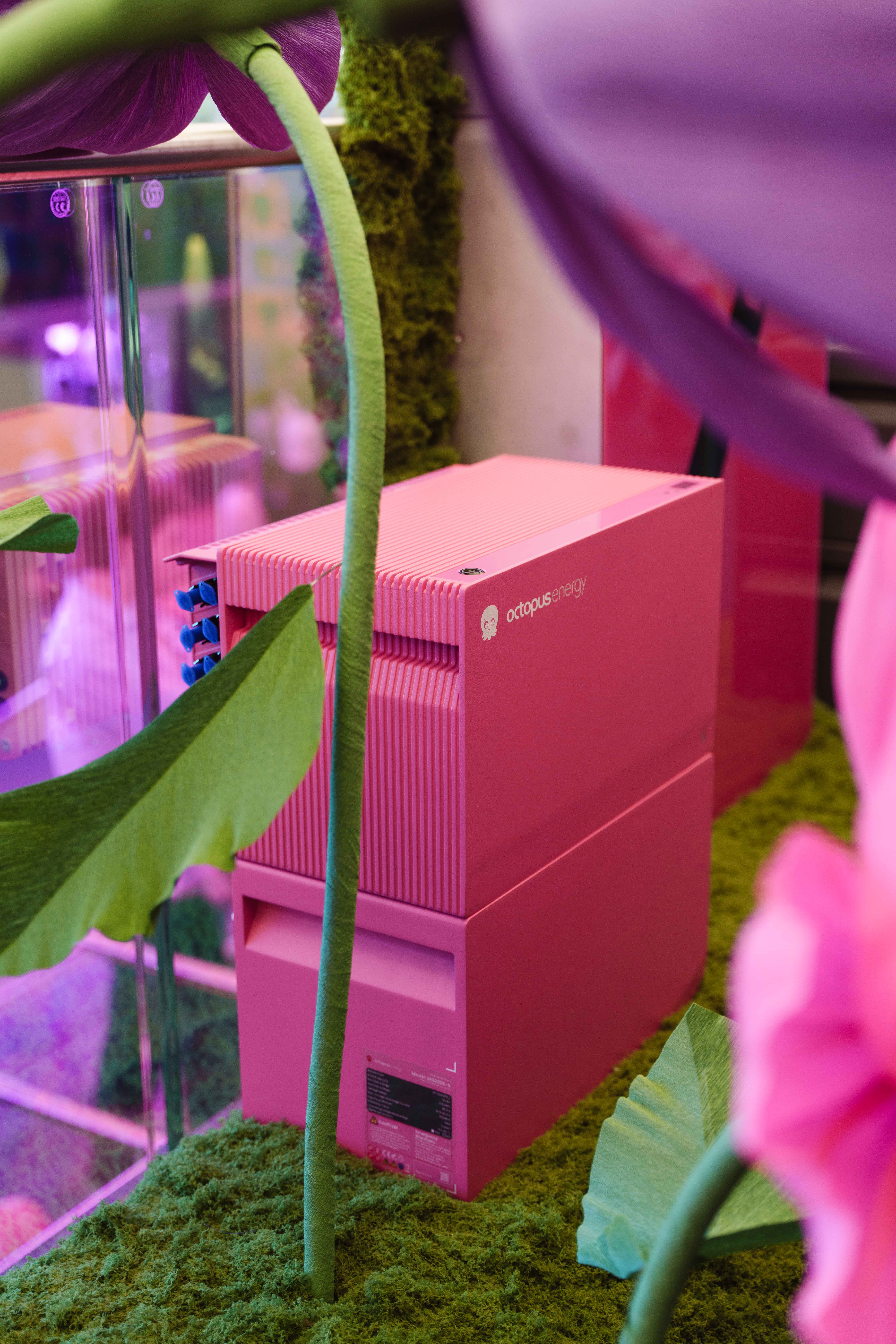 Pink electronic device in a lush green setting, surrounded by artificial plants under purple lighting.