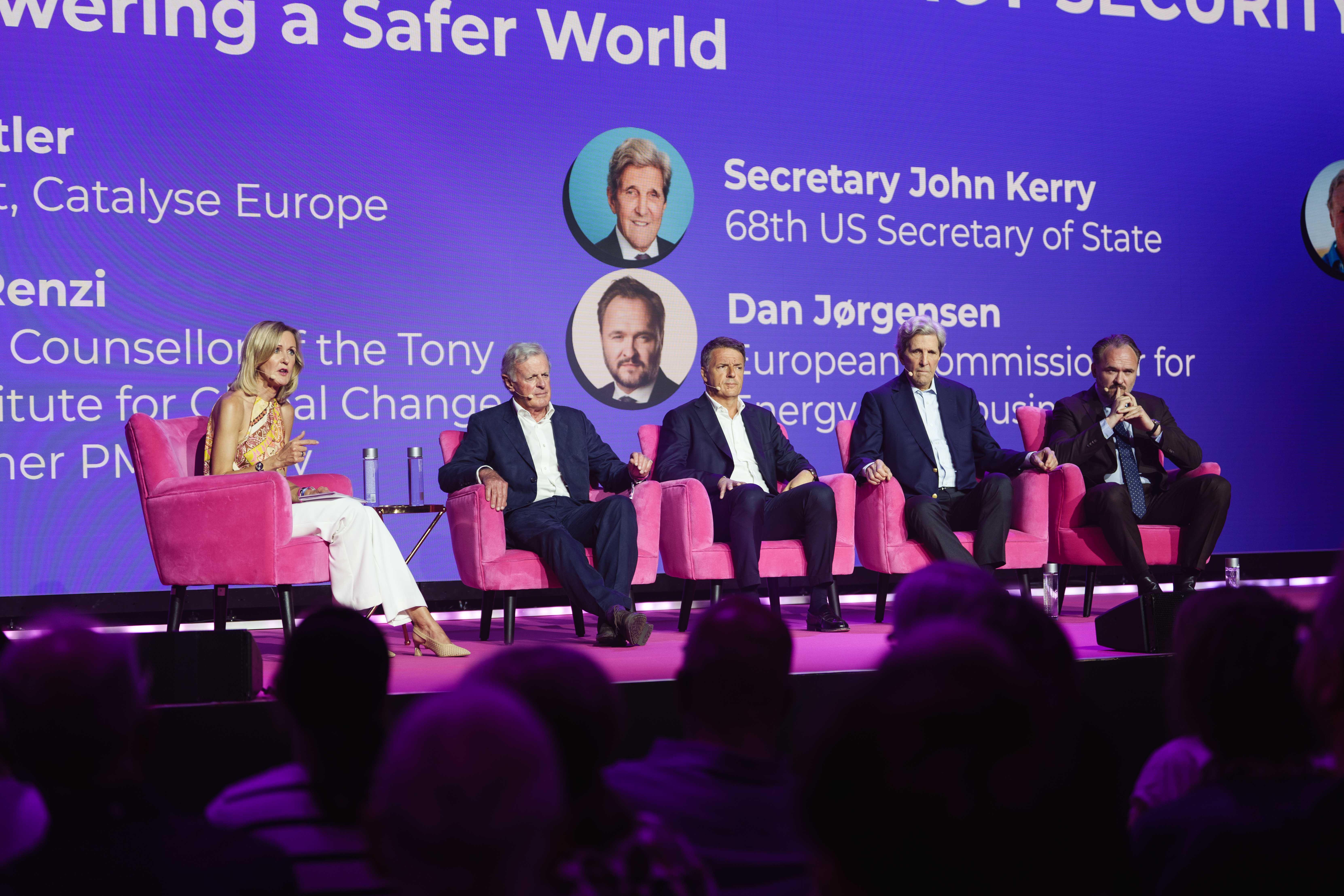 A panel of five people in pink chairs on stage, with a purple backdrop displaying names and titles, addressing an audience.