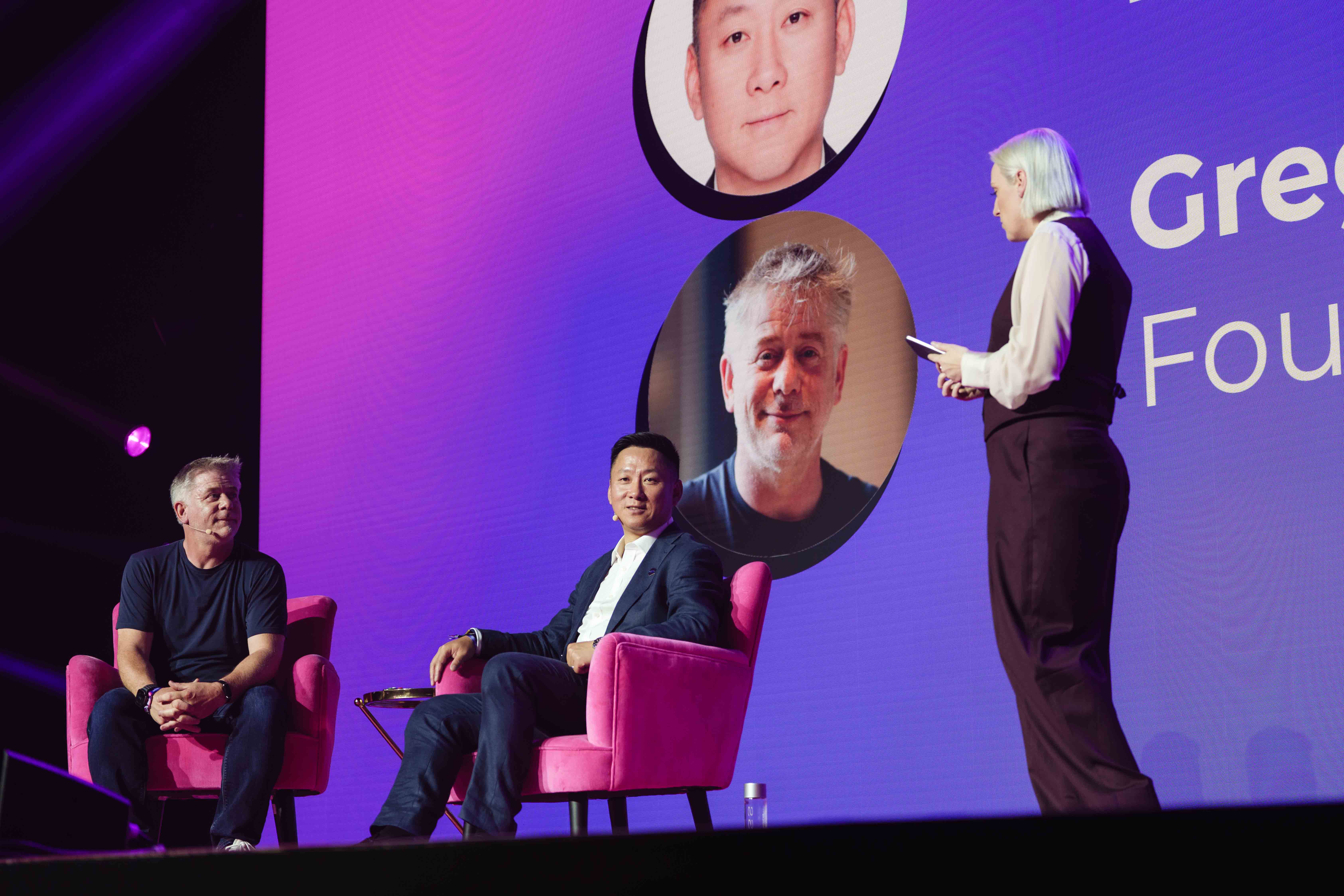 Three people on stage engage in a panel discussion, with a large screen displaying headshots and text in the background.