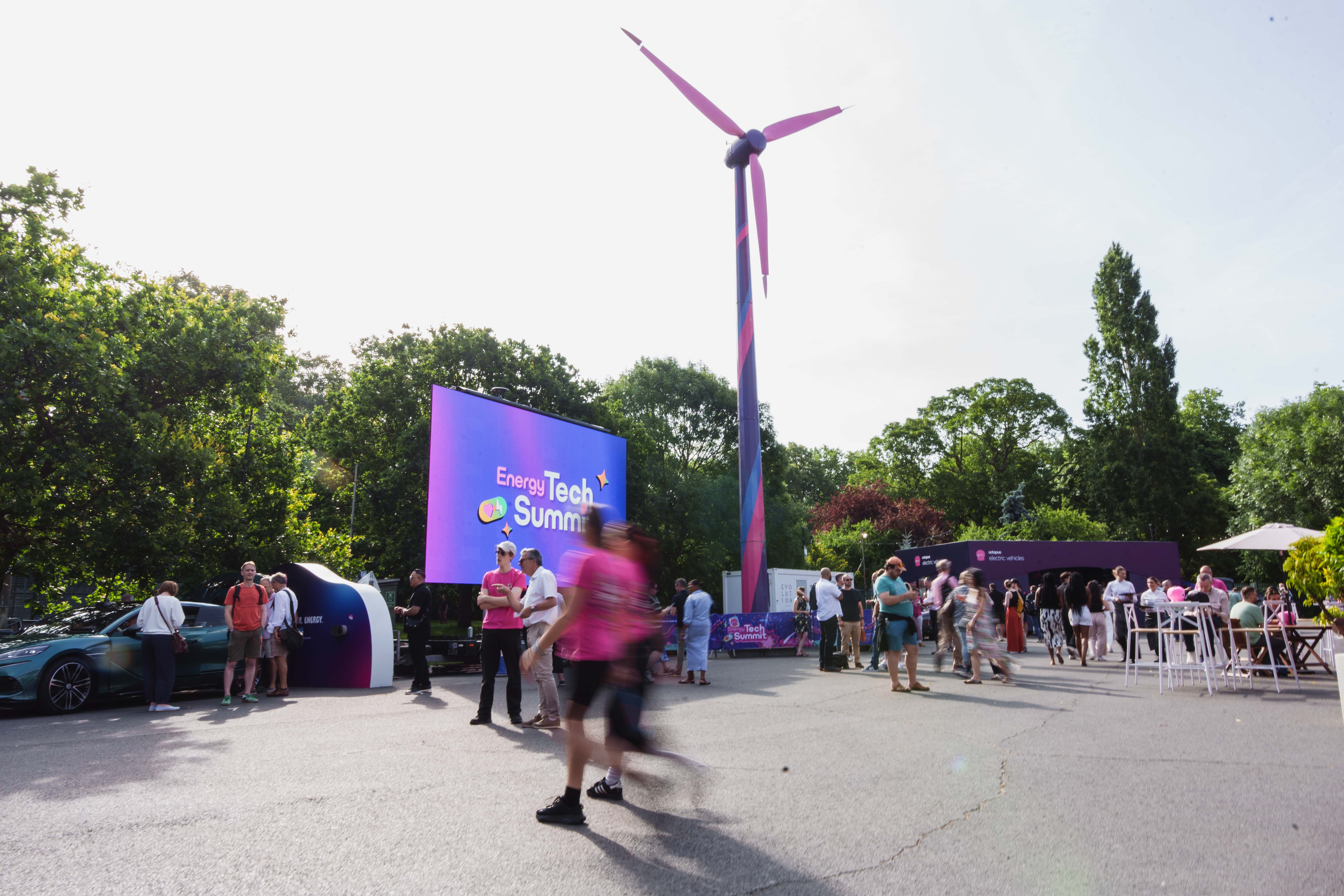 Outdoor event scene with people, a large screen displaying "Energy Tech Summit," and a tall, colorful wind turbine surrounded by trees.