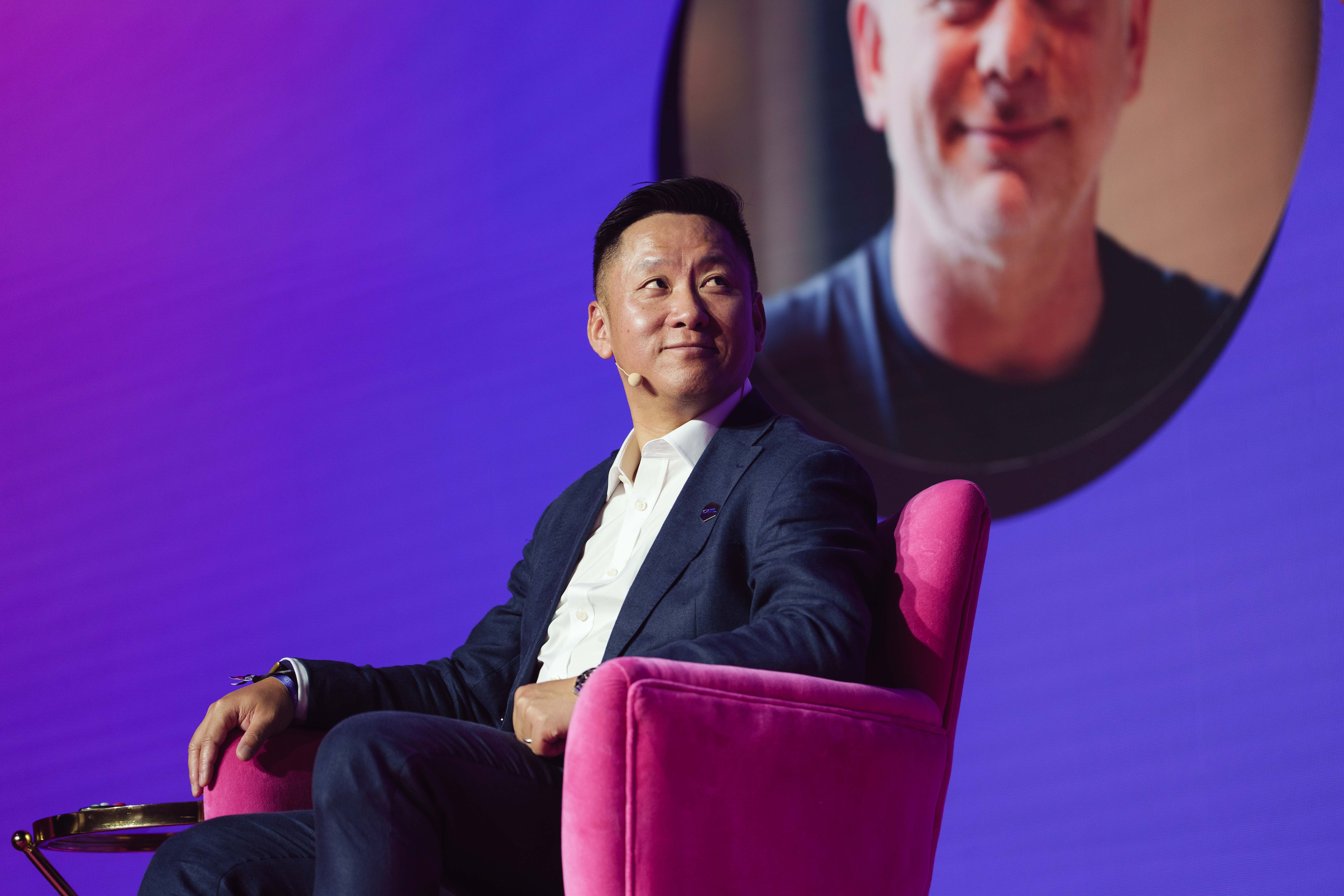 A person in a suit sits on a pink chair at an event, with a headset mic, against a purple backdrop featuring a blurred portrait.