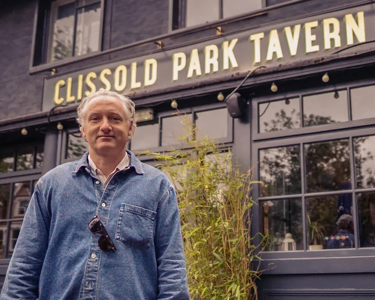 A person in a denim shirt stands in front of Clissold Park Tavern, with a bamboo plant and large windows visible in the background.