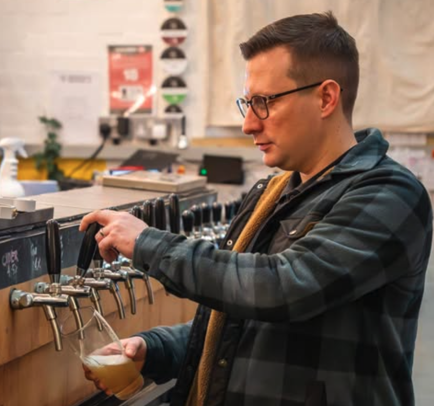 A person in a checkered jacket pours a drink from a tap handle into a glass at a bar with multiple taps.