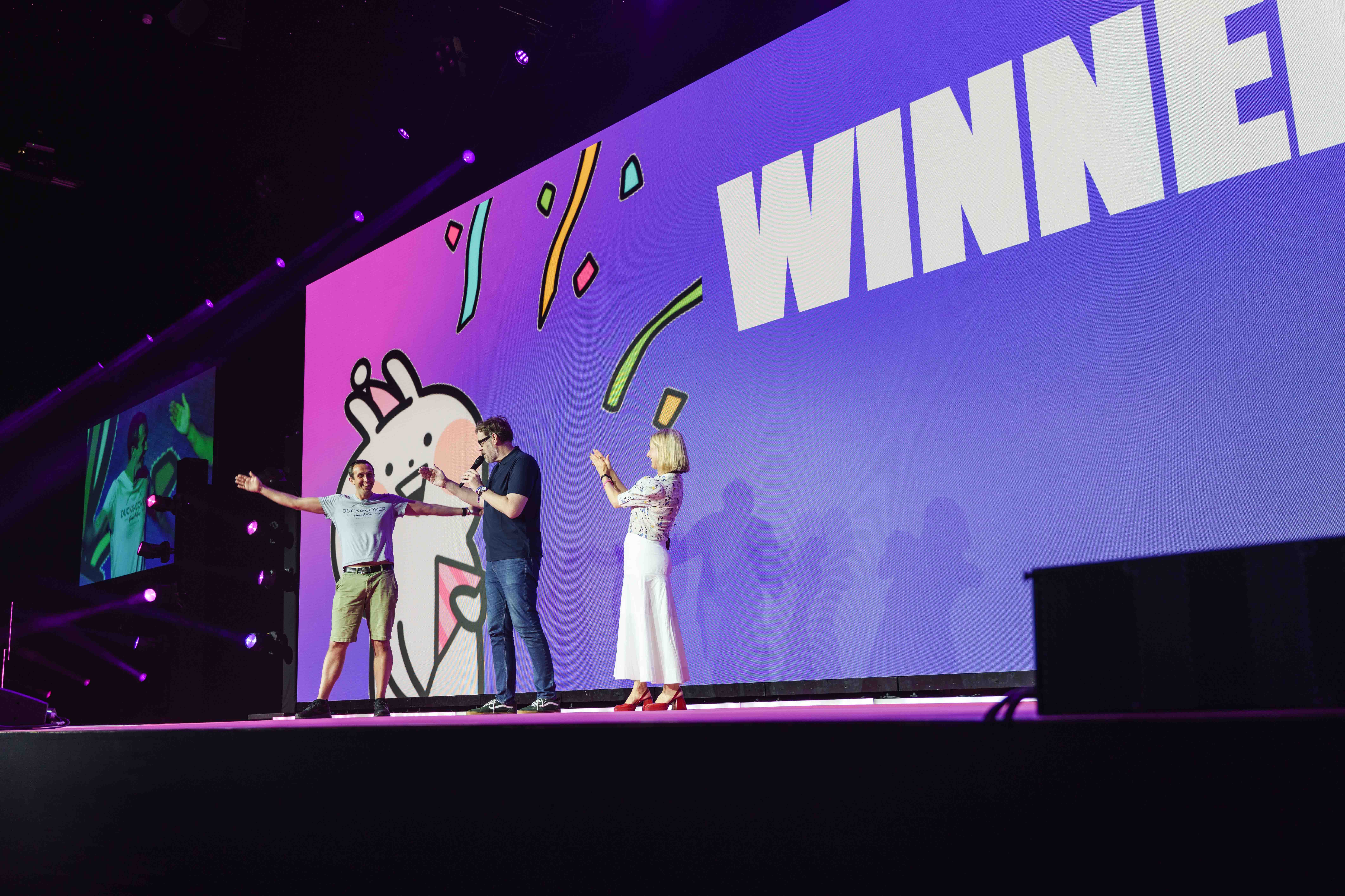 Three people on stage celebrate a winner, with a large screen displaying "WINNER" and festive graphics in the background.