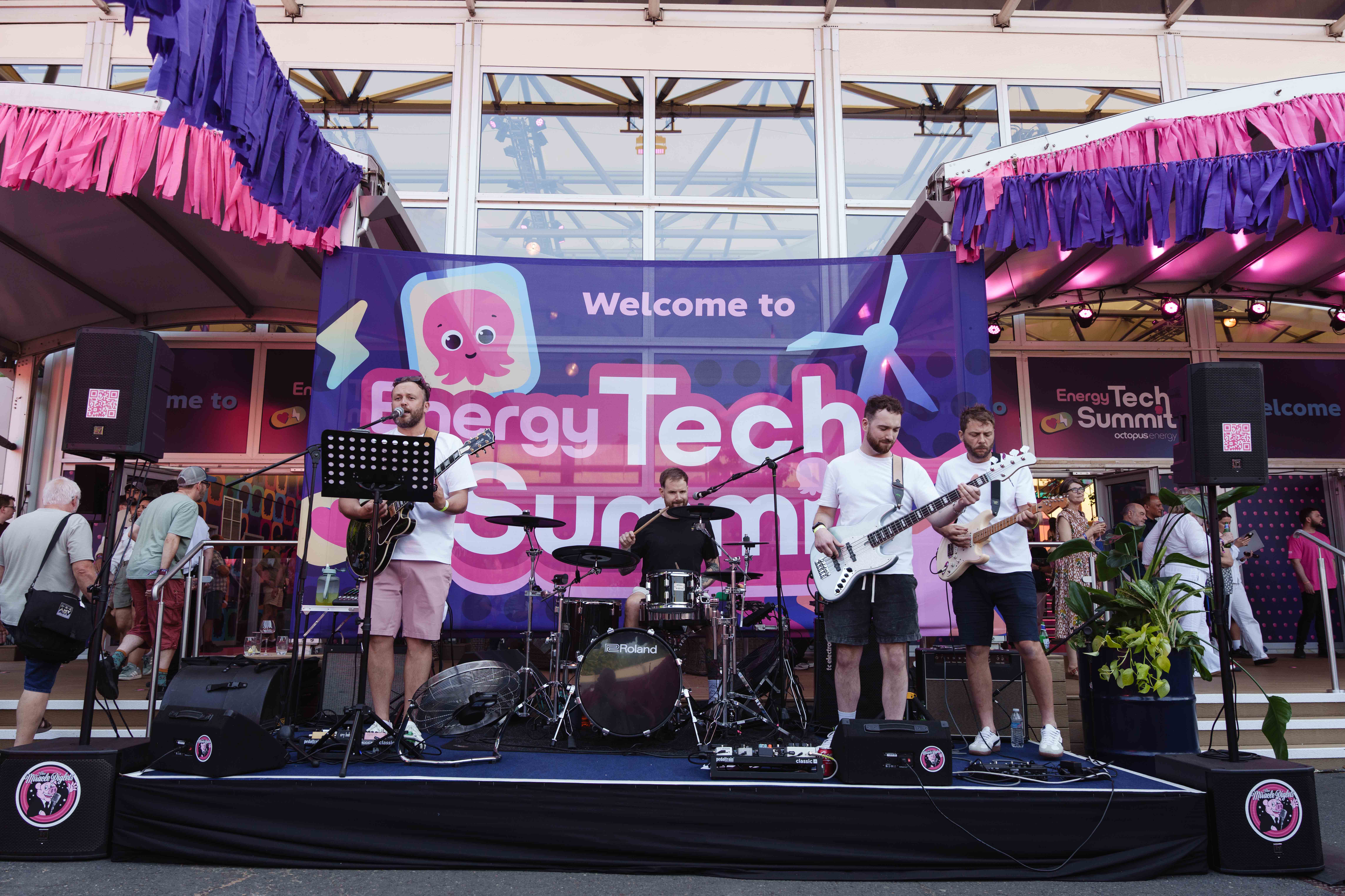 A band performs on stage at the Energy Tech Summit, with vibrant banners and decorations, surrounded by attendees.