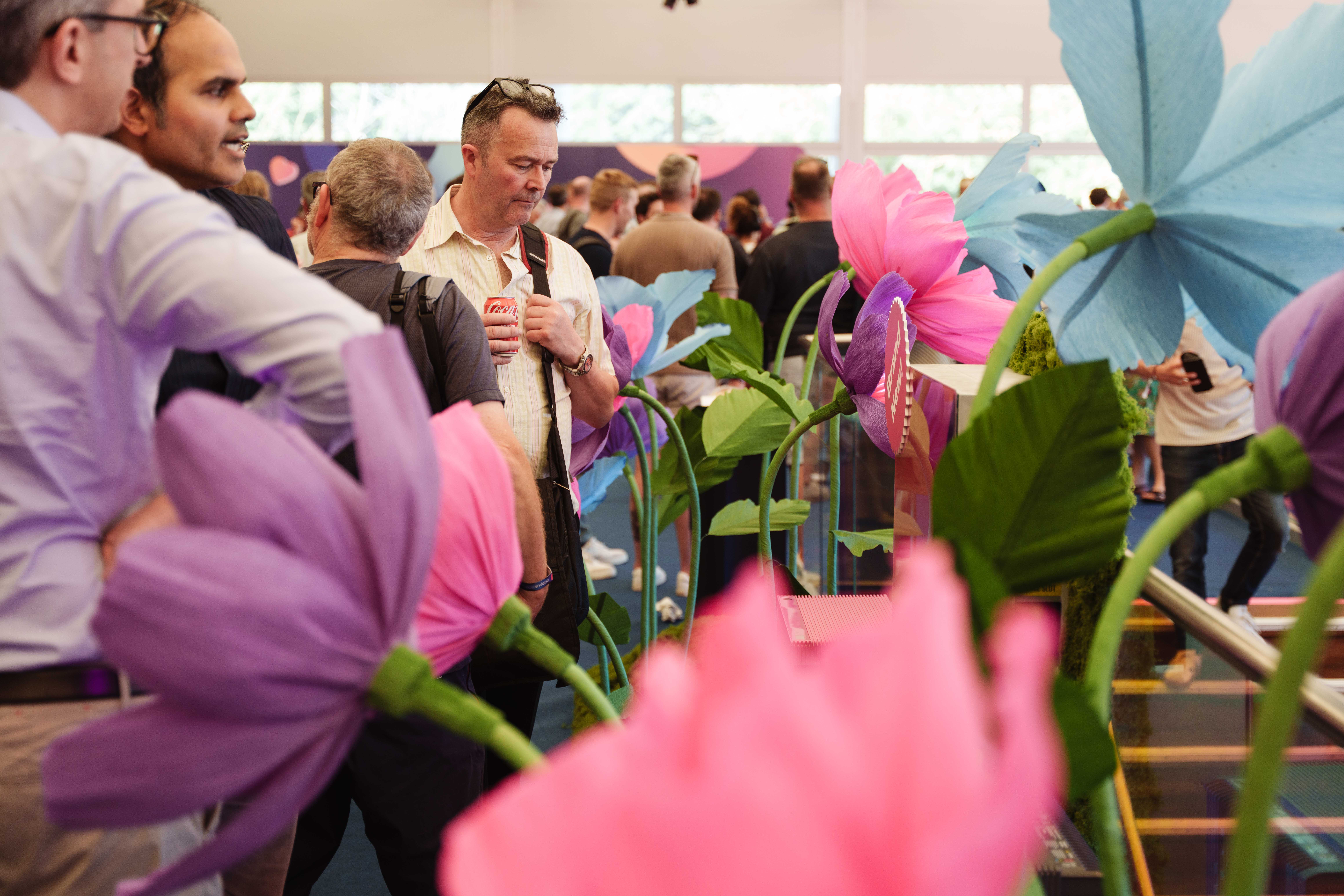People gathered around colorful, oversized flowers in an indoor event space, with a man holding a drink can in the foreground.