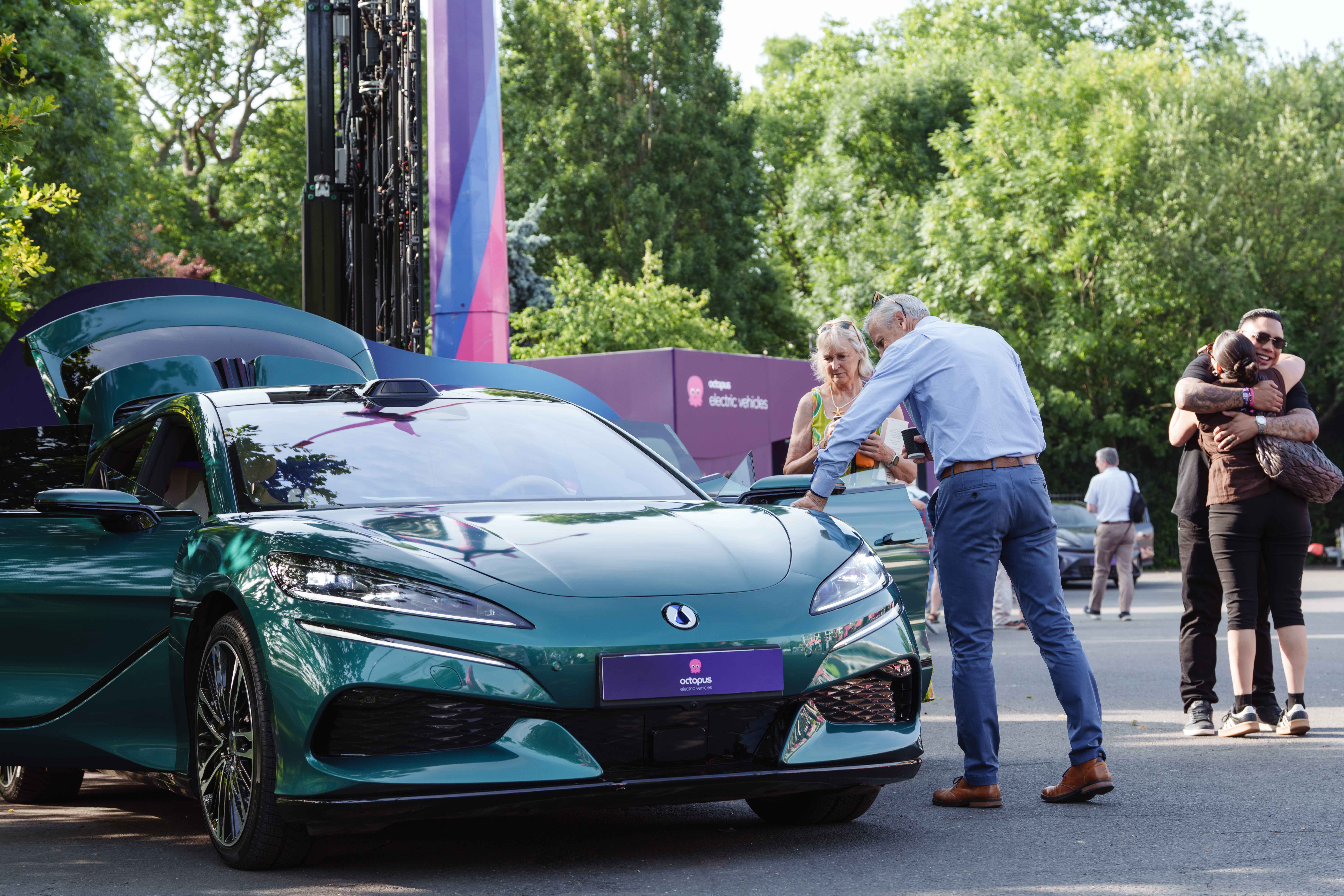 A sleek green car is on display outdoors. A man and woman examine it while two people hug nearby. Trees and event signage are in the background.