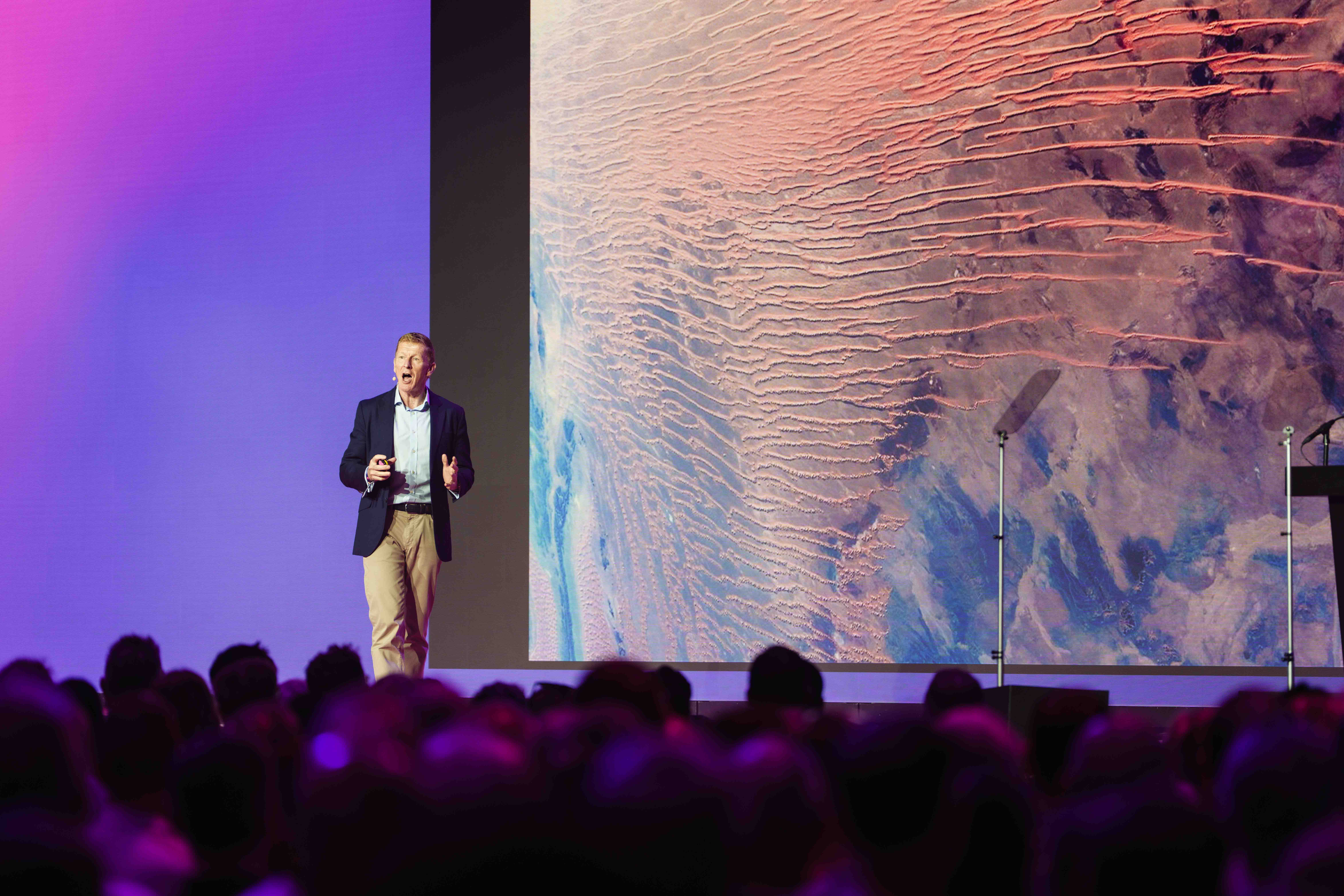 A speaker on stage gestures during a presentation with a vivid, abstract image displayed on a large screen behind them.