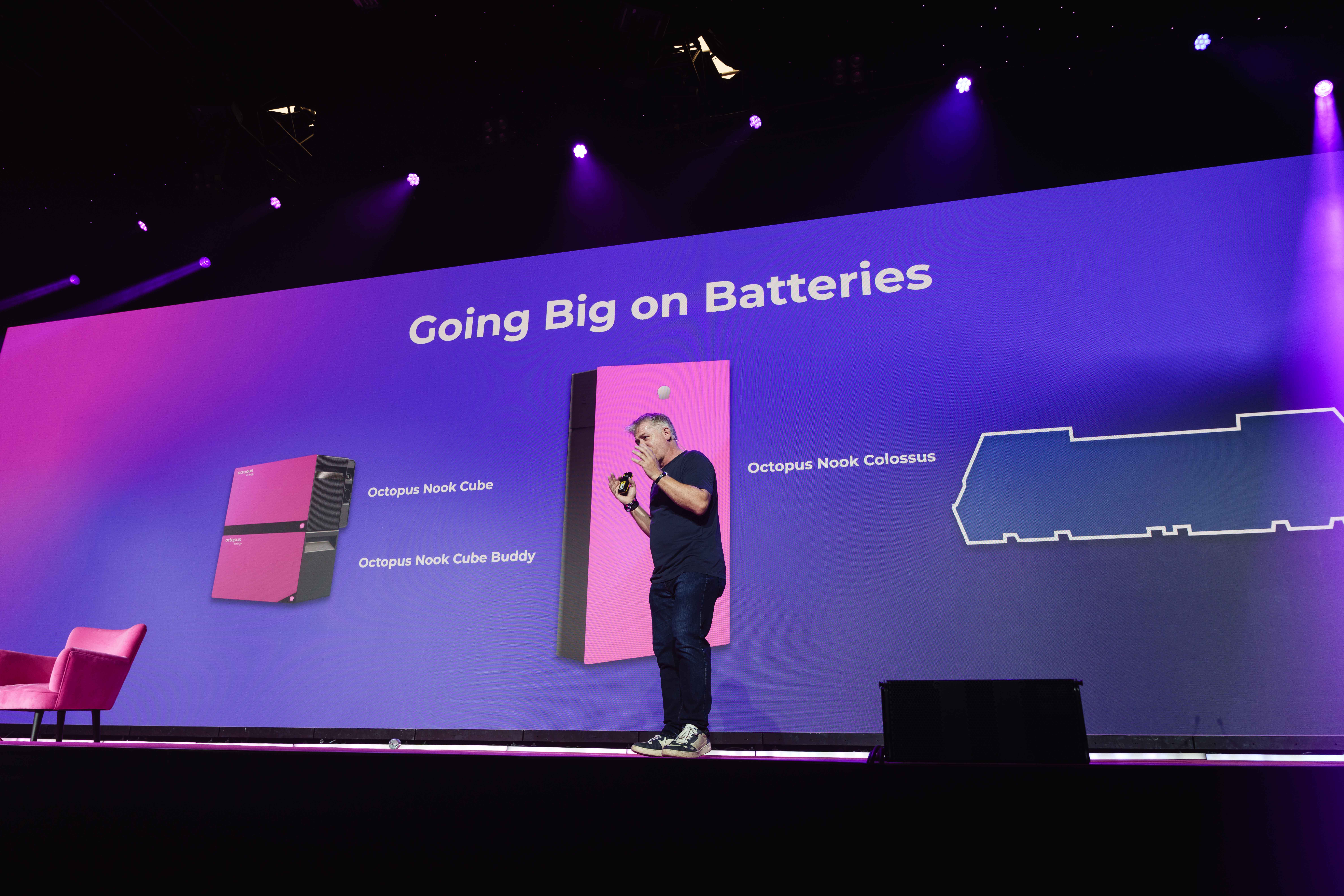 Man on stage presenting "Going Big on Batteries" with visuals of Octopus Nook Cube and Colossus equipment against a purple backdrop.