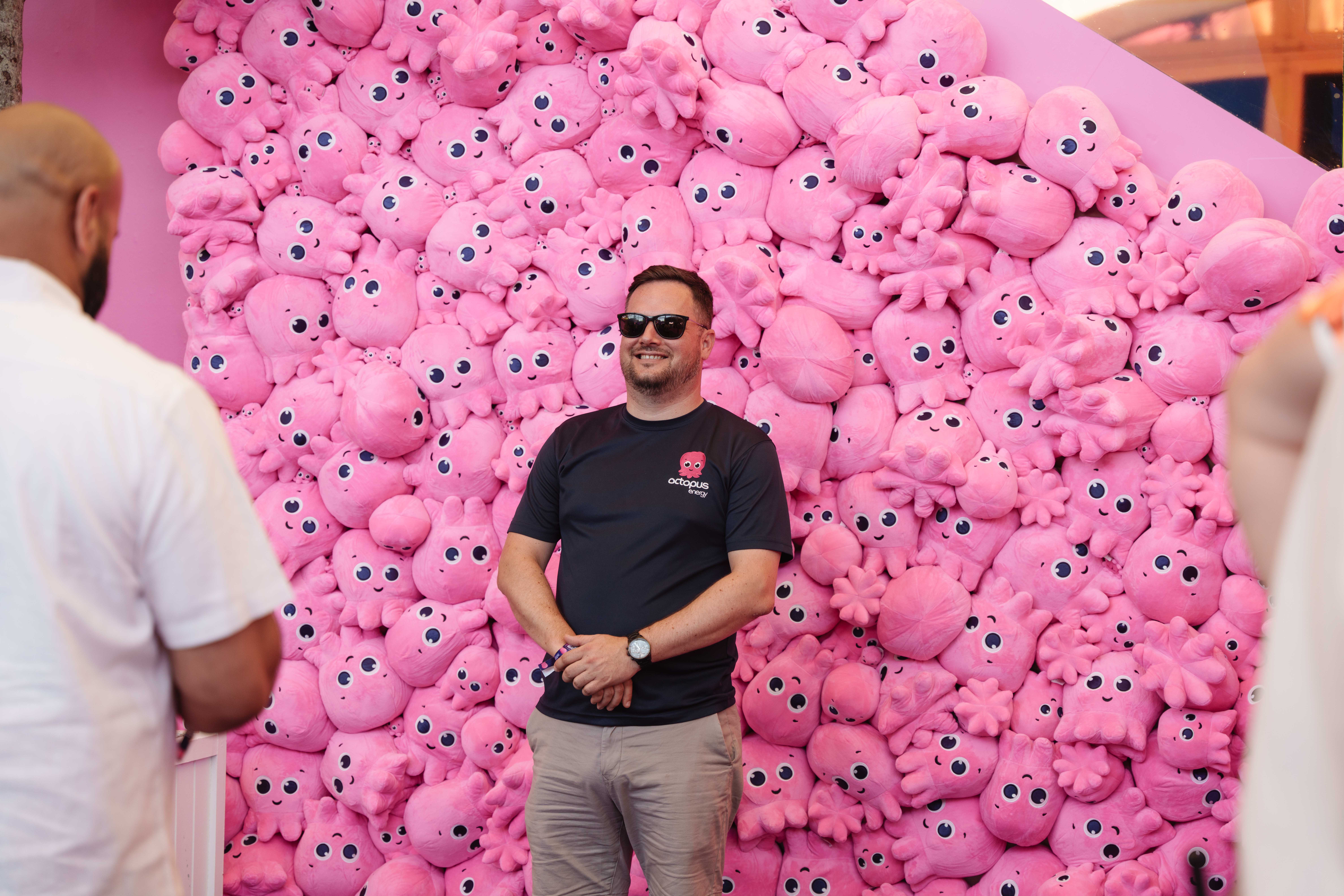 Man in sunglasses stands before a wall of pink octopus plushies, smiling and wearing a black shirt with an octopus logo.