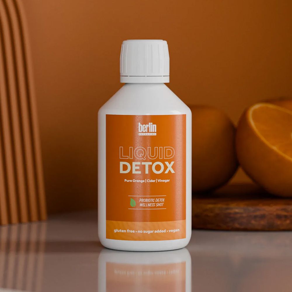 A white plastic bottle with a white screw cap labeled “Liquid Detox” in bold orange text, placed on a reflective surface. The label includes details such as “Pure Orange | Cider | Vinegar,” “Promoting Detox Wellness Shot,” and notes that it is gluten-free, sugar-free, and vegan. The background features an orange color scheme with whole and halved oranges on a wooden board, complementing the product theme.