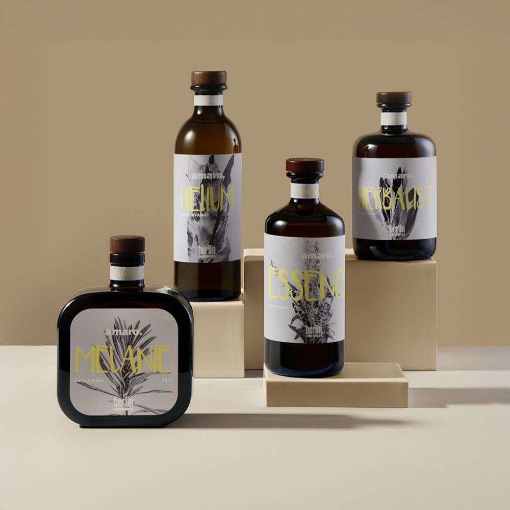 Bitter Mania collection by Berlin Packaging featuring Helium, Essence, Herbalist and Melanie glass bottles for bitters and distilled spirits, showcasing distinctive bottle shapes, dark glass finishes, wooden closures and coordinated label designs on a neutral display setting.