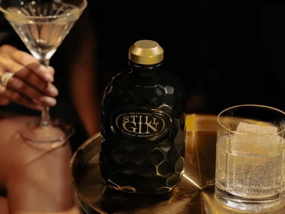 A dark, textured bottle of gin on a table next to a glass with ice and a person's hand holding a cocktail glass.