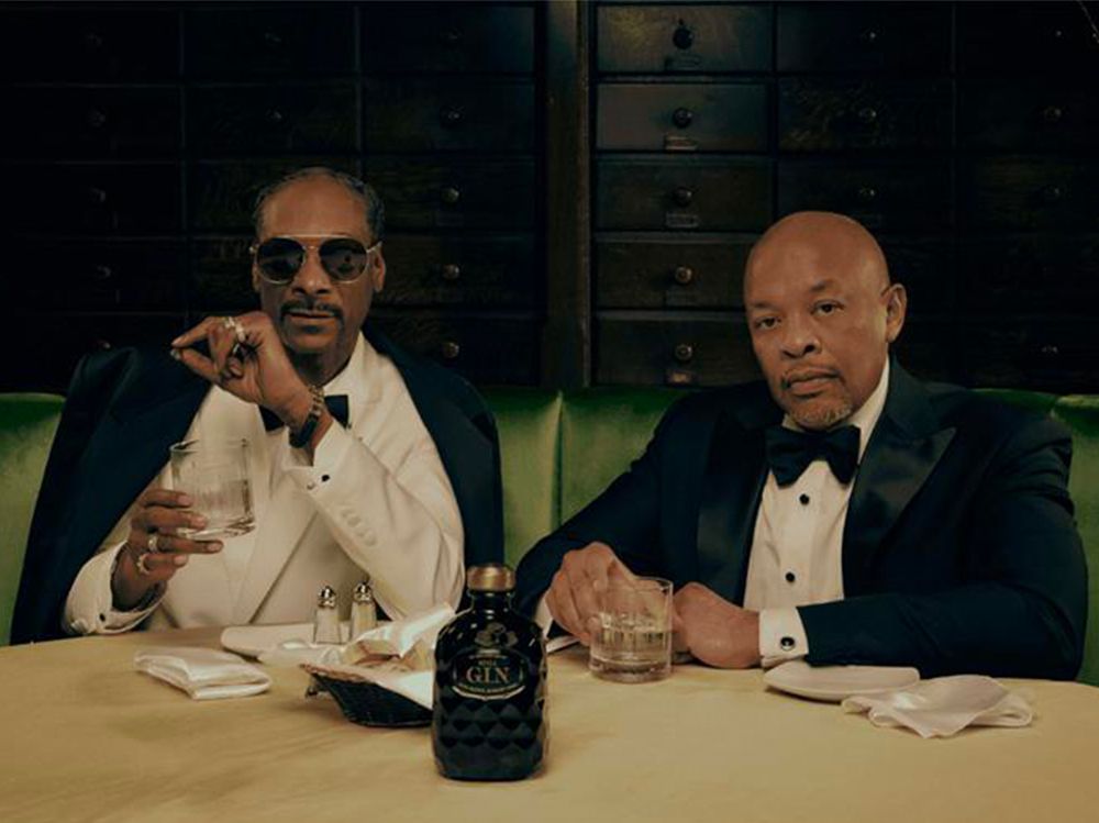 Two men in tuxedos sit at a table with drinks, one holding a cigar. A bottle of gin is on the table, and a dark wooden backdrop is behind them.