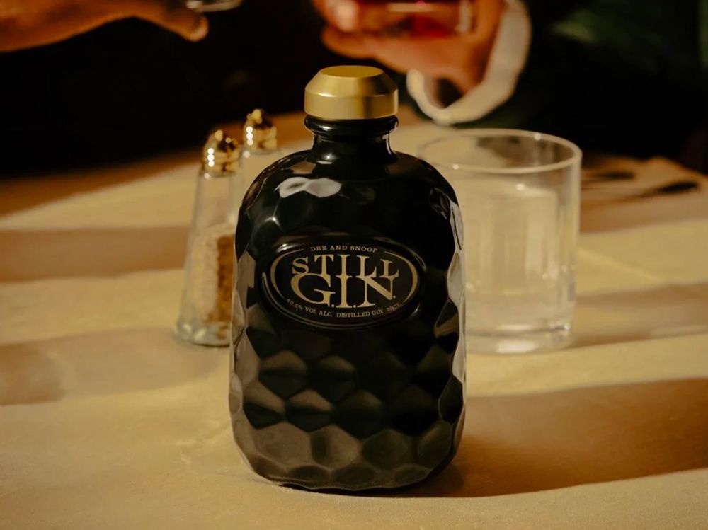 A dark, textured bottle of Stilk Gin sits on a table with a glass, pepper shaker, and blurred individuals in the background.
