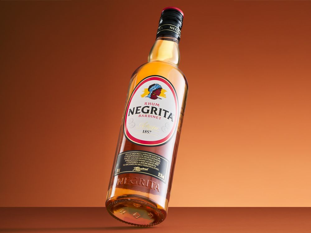 A bottle of Negrita Bardinet rhum is balanced at an angle against an orange background. The label features the brand name and a colorful logo.