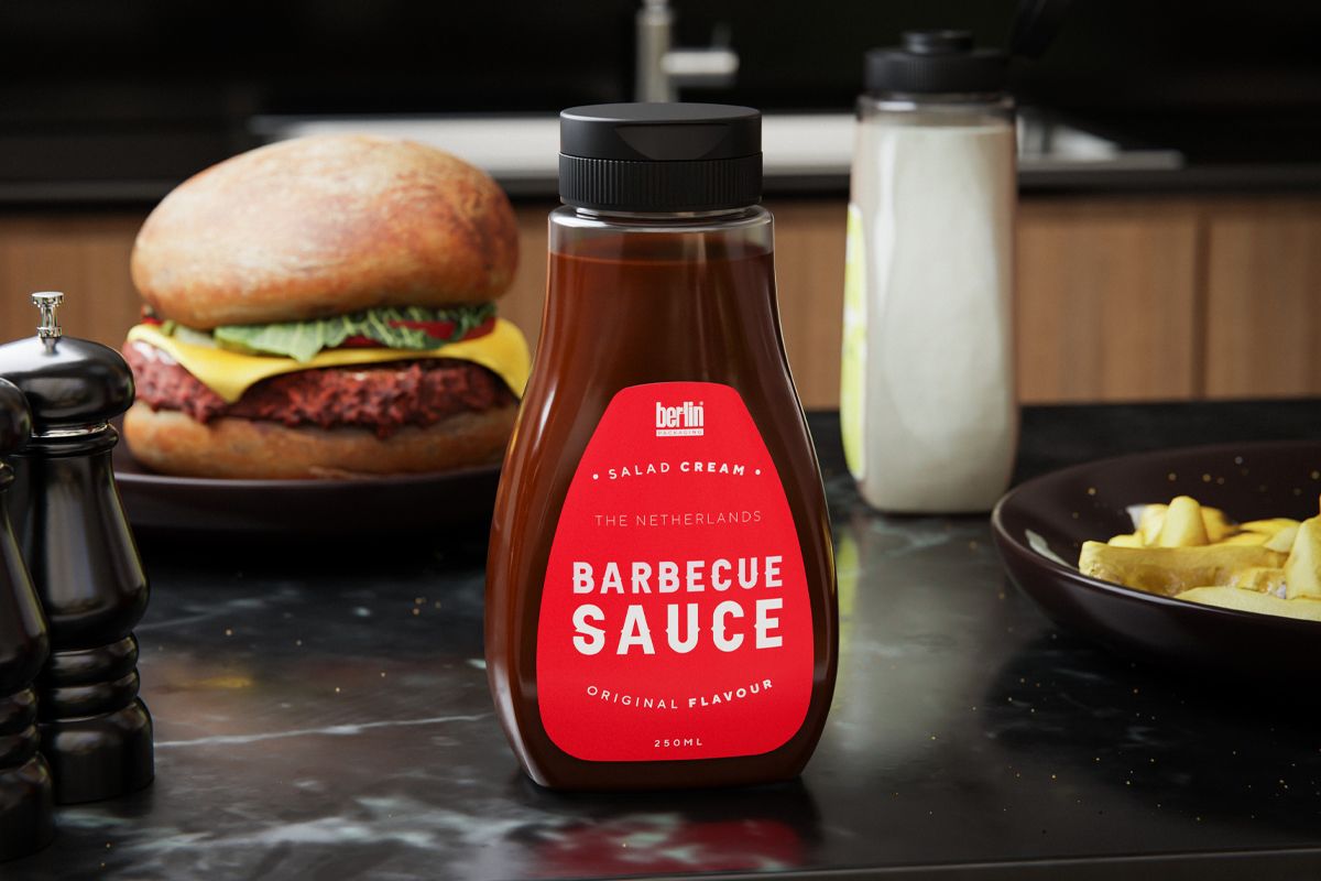 A bottle of “Barbecue Sauce” labeled “The Netherlands” and “Original Flavour” stands on a dark countertop. In the background, there is a burger with lettuce and cheese, a bottle of sauce, and a bowl of fries.