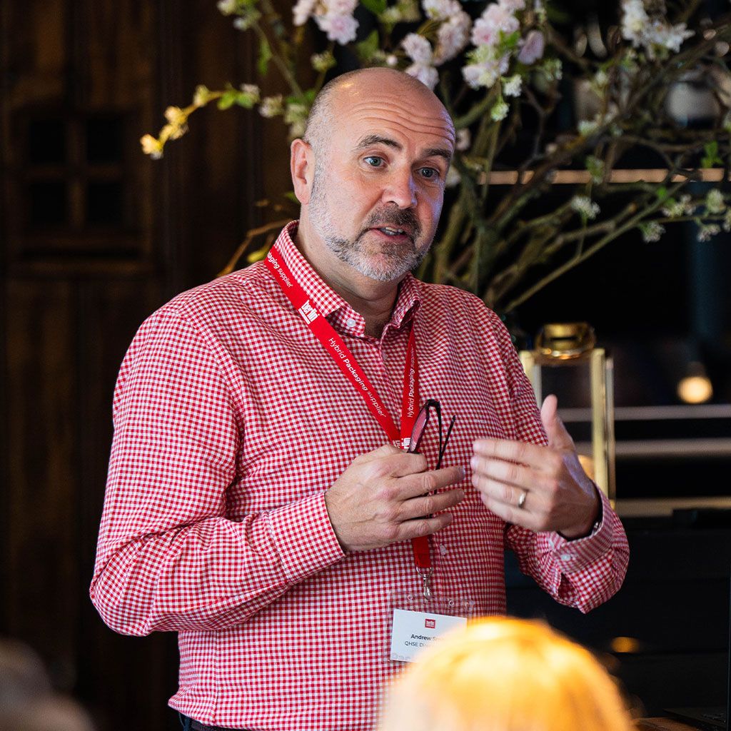 Man in a red checkered shirt speaks, gesturing with his hands, wearing a name badge and red lanyard. Blossoms are visible in the background.