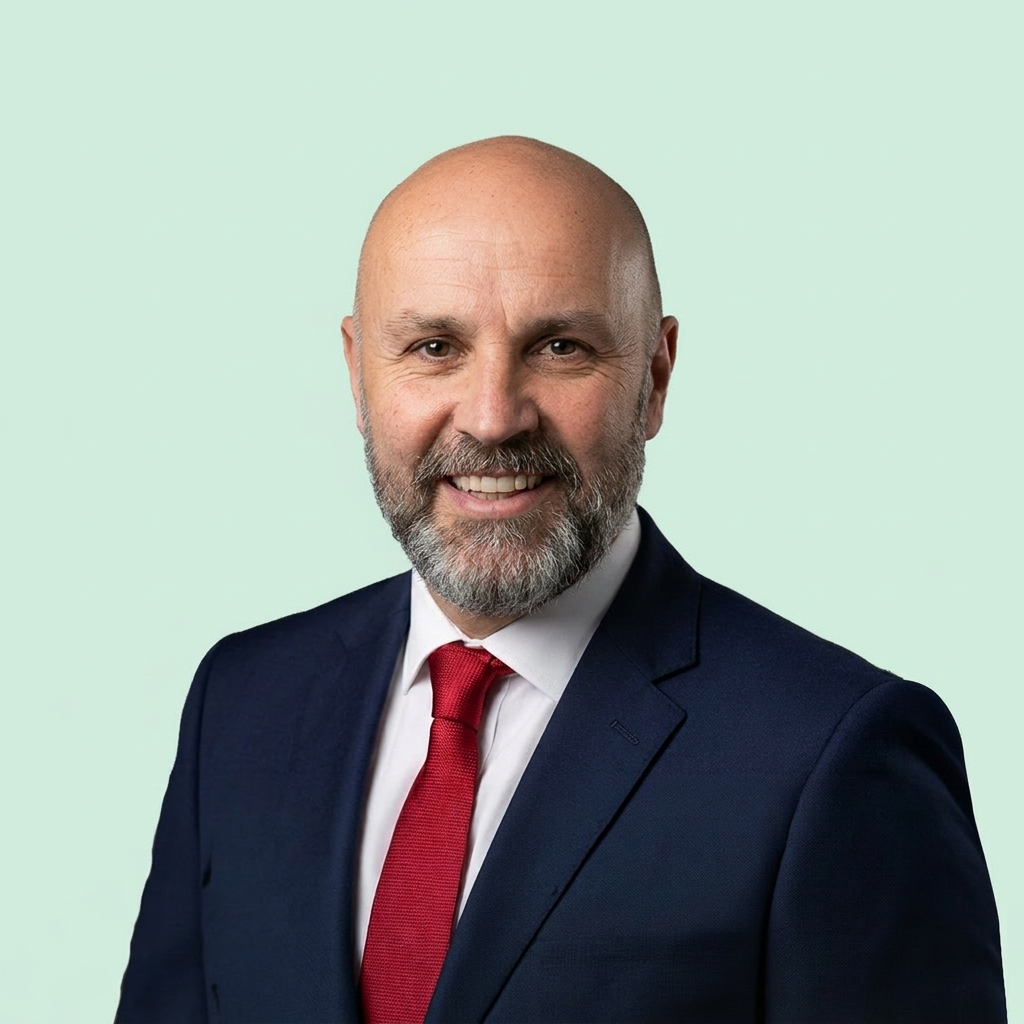Bald man with a beard in a navy suit and red tie, smiling against a light green background.