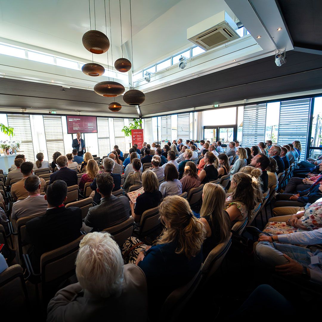 A large group of people seated in a spacious, well-lit conference room with a presenter speaking at the front and a presentation screen visible.