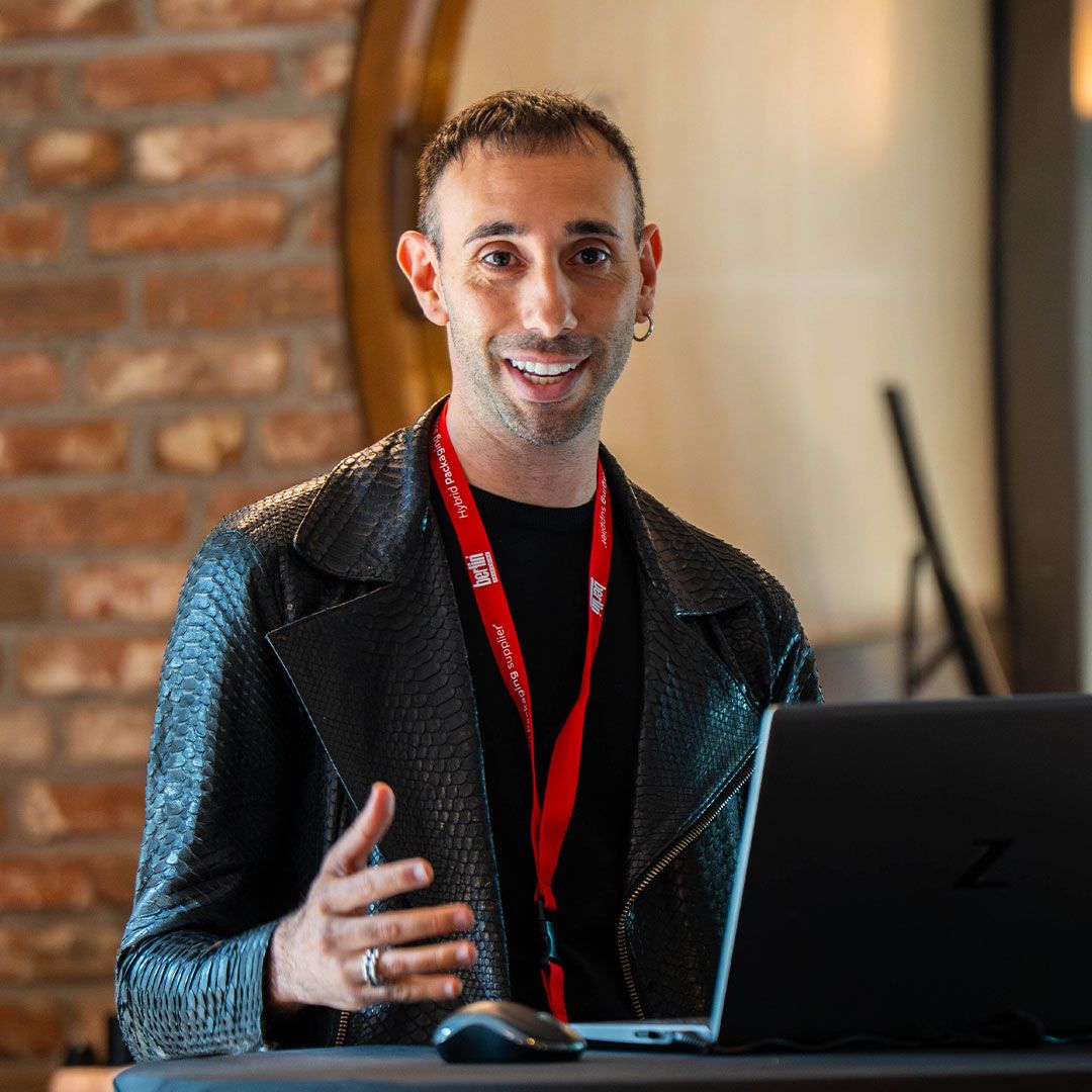 Person with a red lanyard speaking at a podium with a laptop, wearing a black textured jacket, smiling against a brick background.