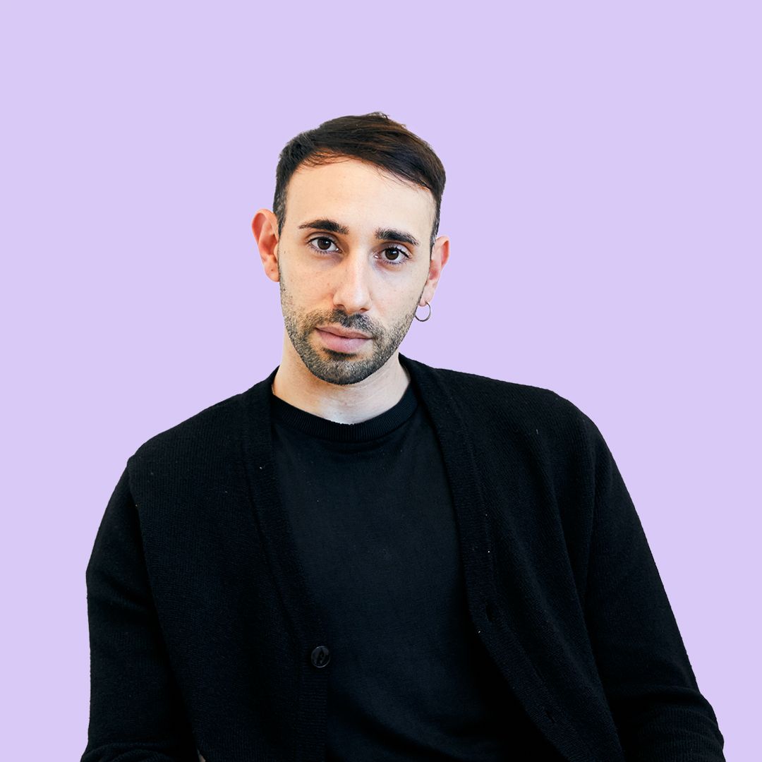 A person with short hair and a hoop earring, wearing a black shirt and cardigan, against a light purple background.