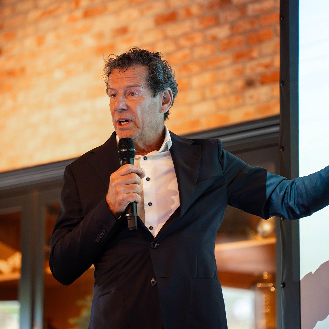 Man in a suit holding a microphone, speaking and gesturing towards a screen, with a brick wall in the background.