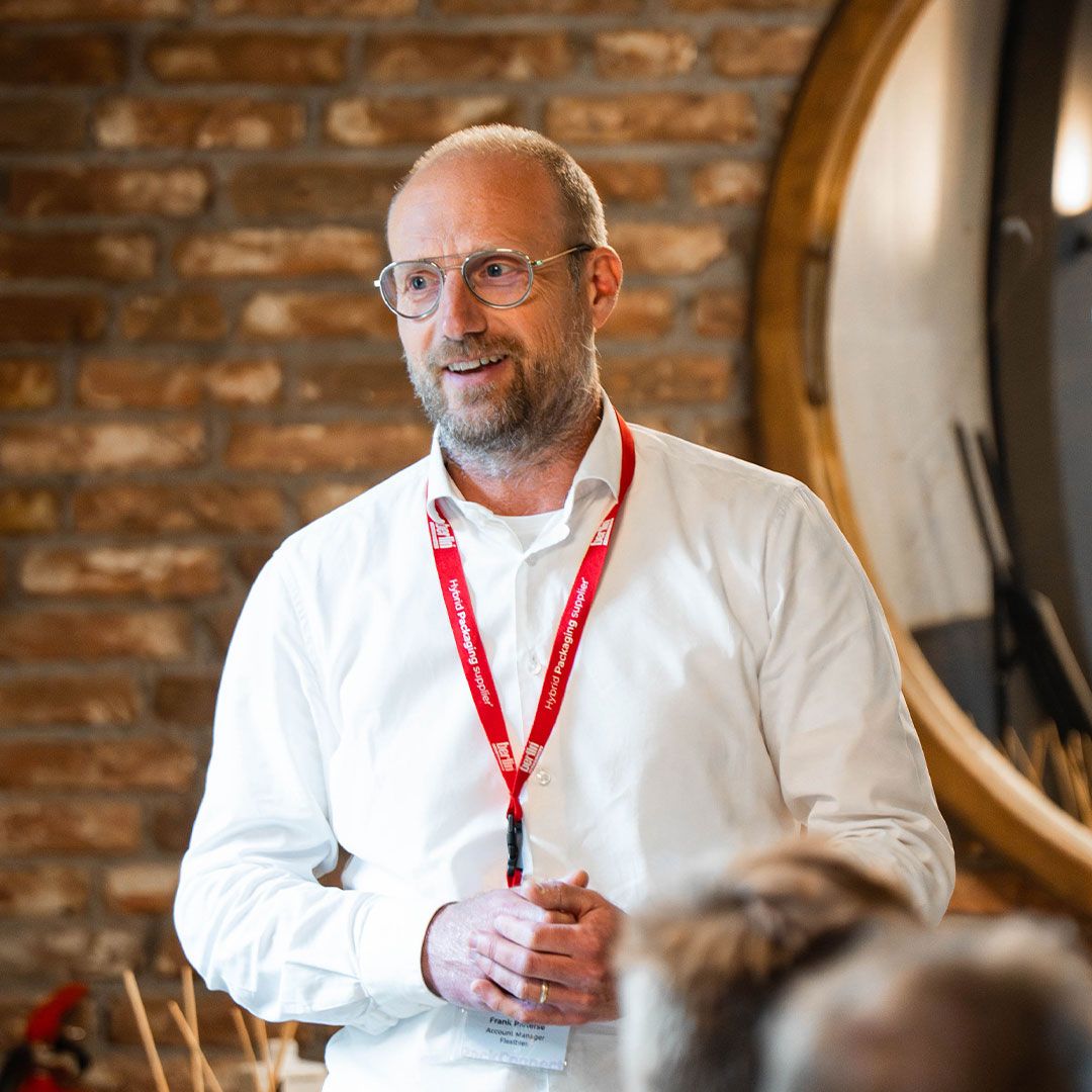 Man with glasses and red lanyard speaks in front of a brick wall, wearing a white shirt, and standing near a large round mirror.